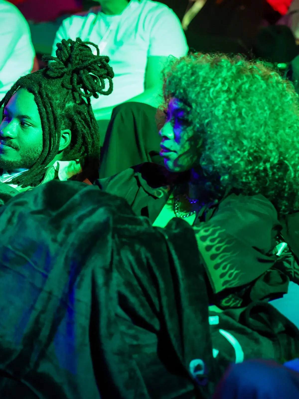 Two people reclining on beanbag seating under saturated green stage light, wrapped in blankets with other attendees visible behind them at a dim, immersive screening event.