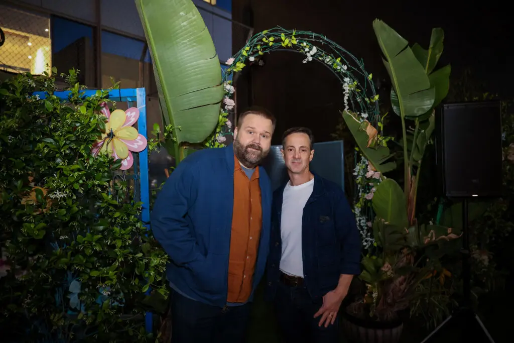 Two guests pose under a green floral arch at a nighttime screening activation, framed by large tropical leaves, potted plants, decorative flowers, and event lighting; one wears an orange shirt and blue jacket.