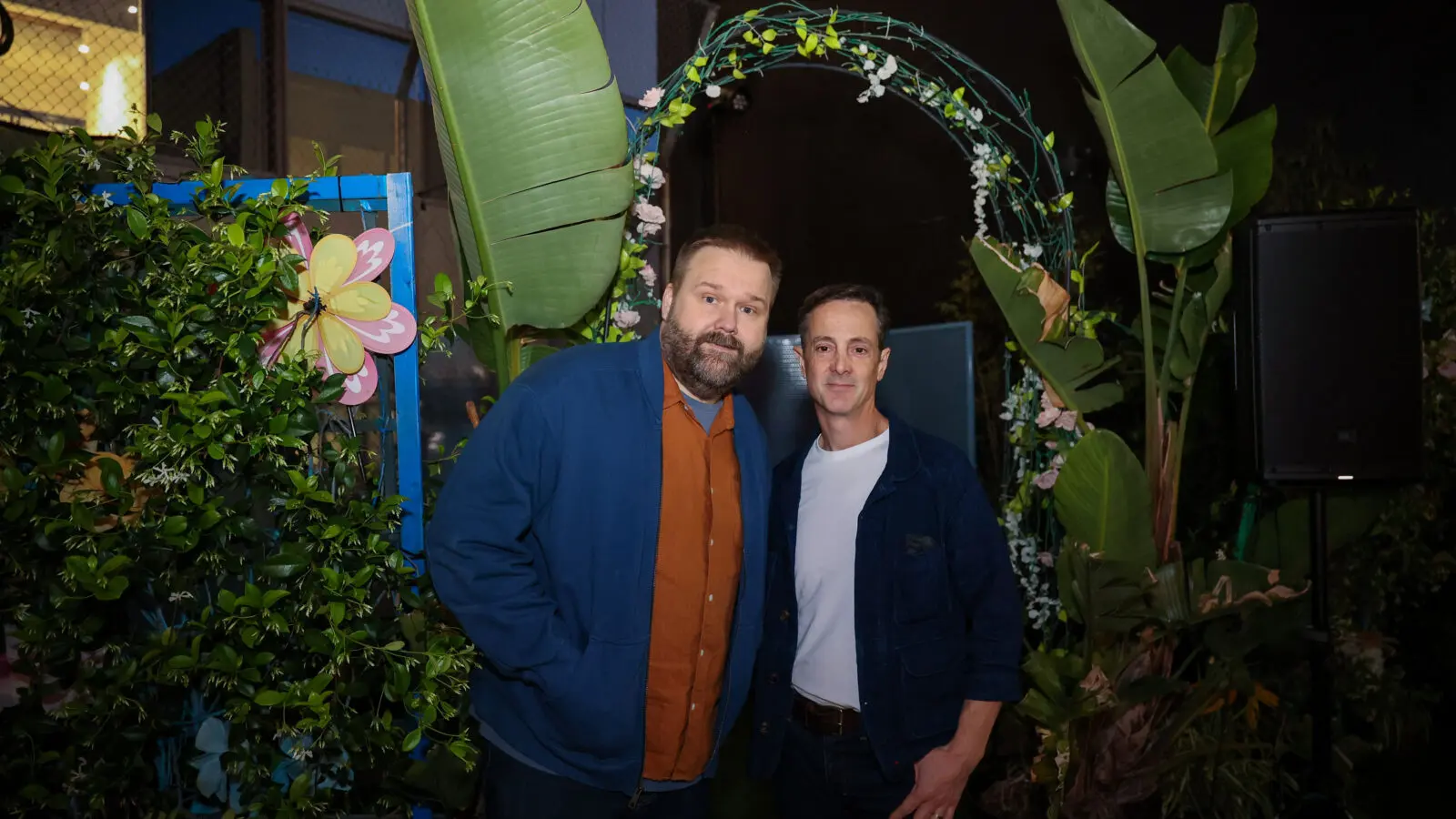 Two men stand under a flowered arch framed by large potted tropical plants and greenery at an outdoor nighttime event.