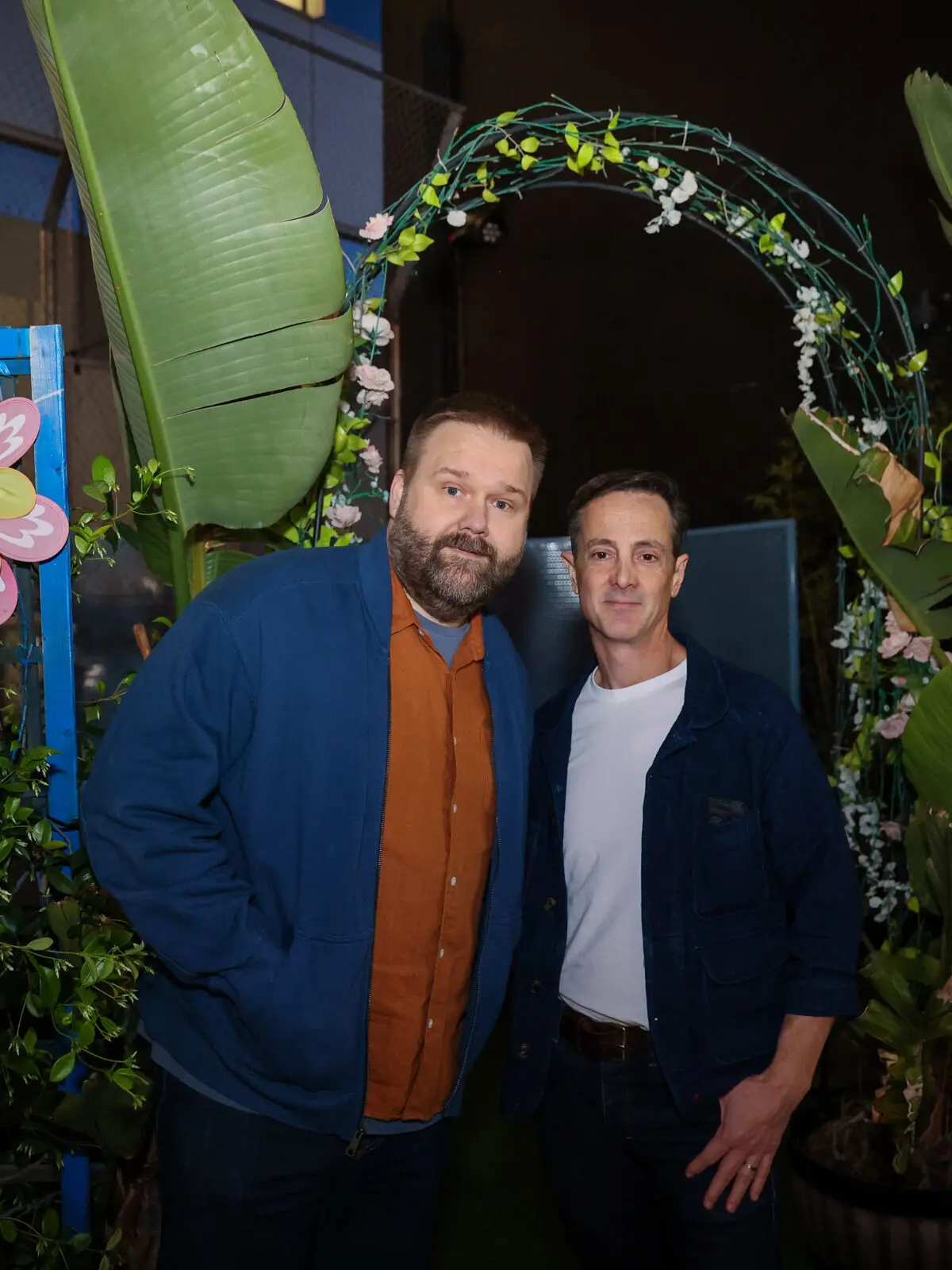 Two men stand under a flowered arch framed by large potted tropical plants and greenery at an outdoor nighttime event.