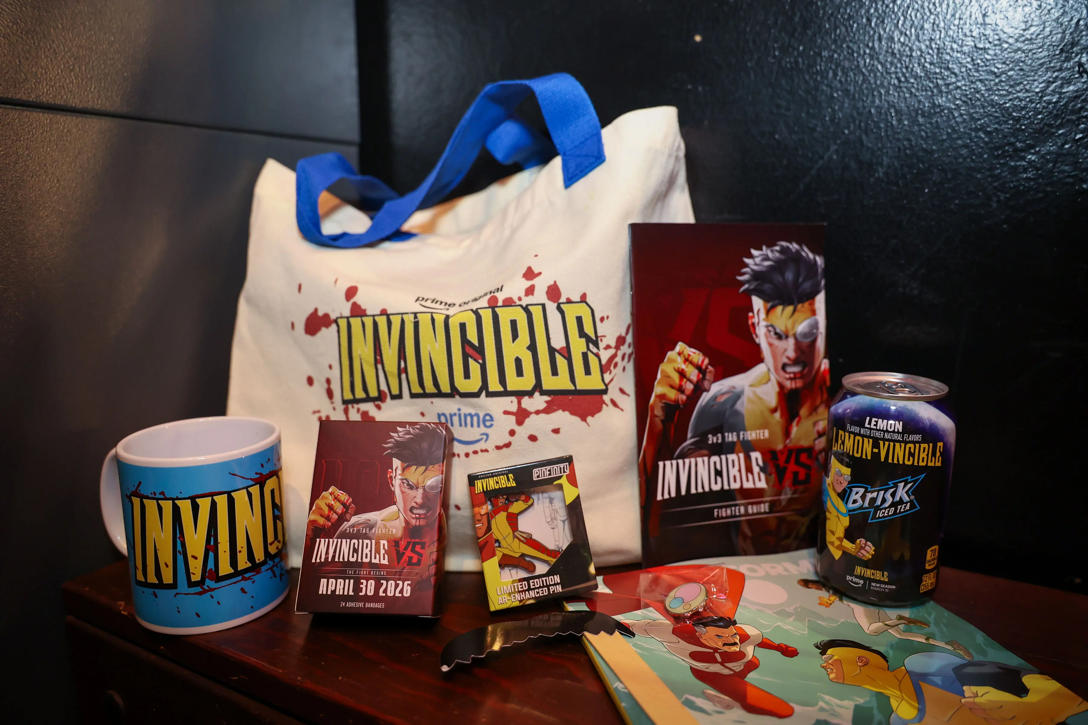 Canvas tote with yellow-and-blue 'Invincible' logo and blood-splatter print, matched by a branded mug, fighter guidebooks, enamel pin, playing cards, and a Brisk 'Lemon-Vincible' can on a wooden table.