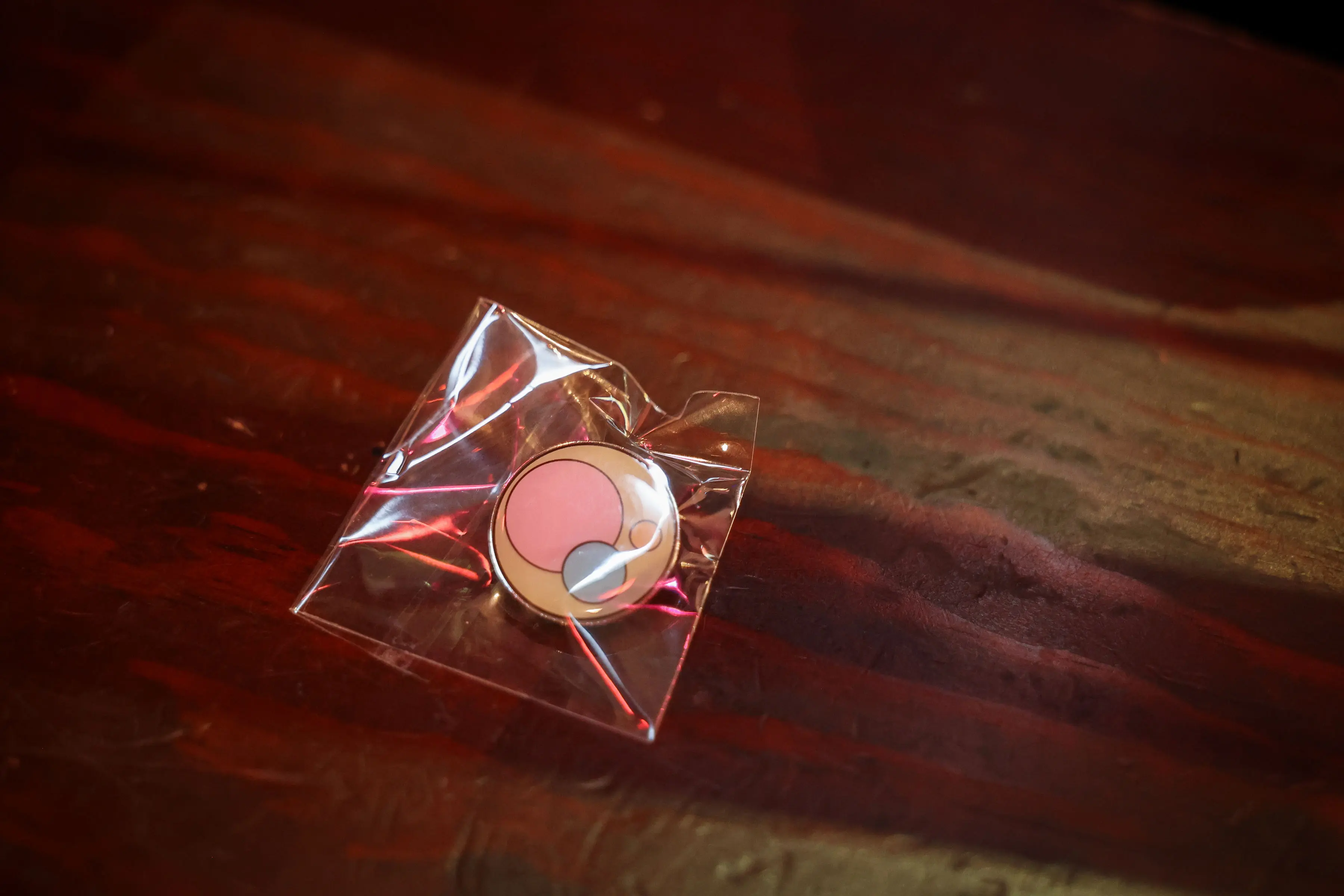 A round enamel pin with pink, cream, and white abstract shapes sits inside clear wrapping on a dark, textured surface.