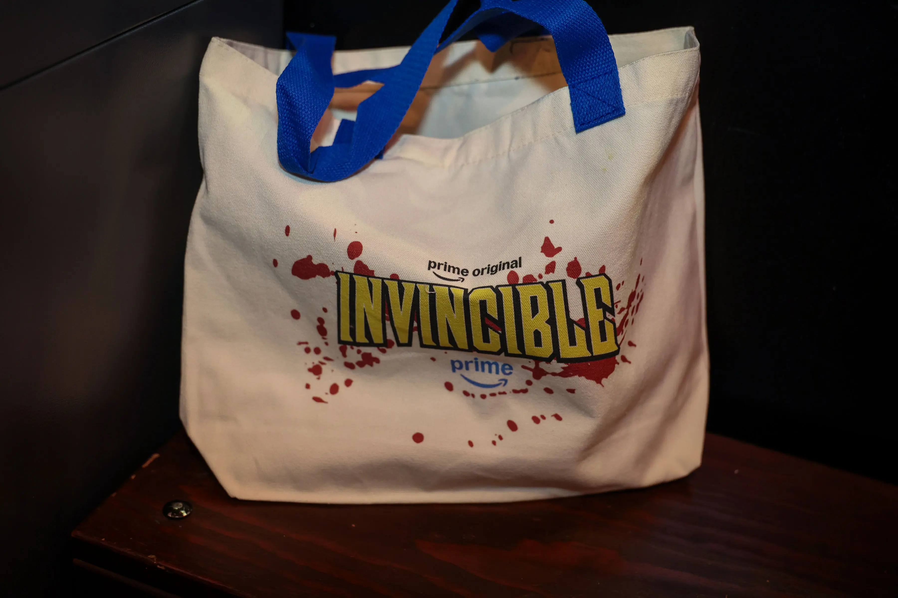 Cream canvas Invincible tote bag with blue handles, yellow logo artwork, Prime branding, and red blood-spatter graphics displayed on a wooden surface.