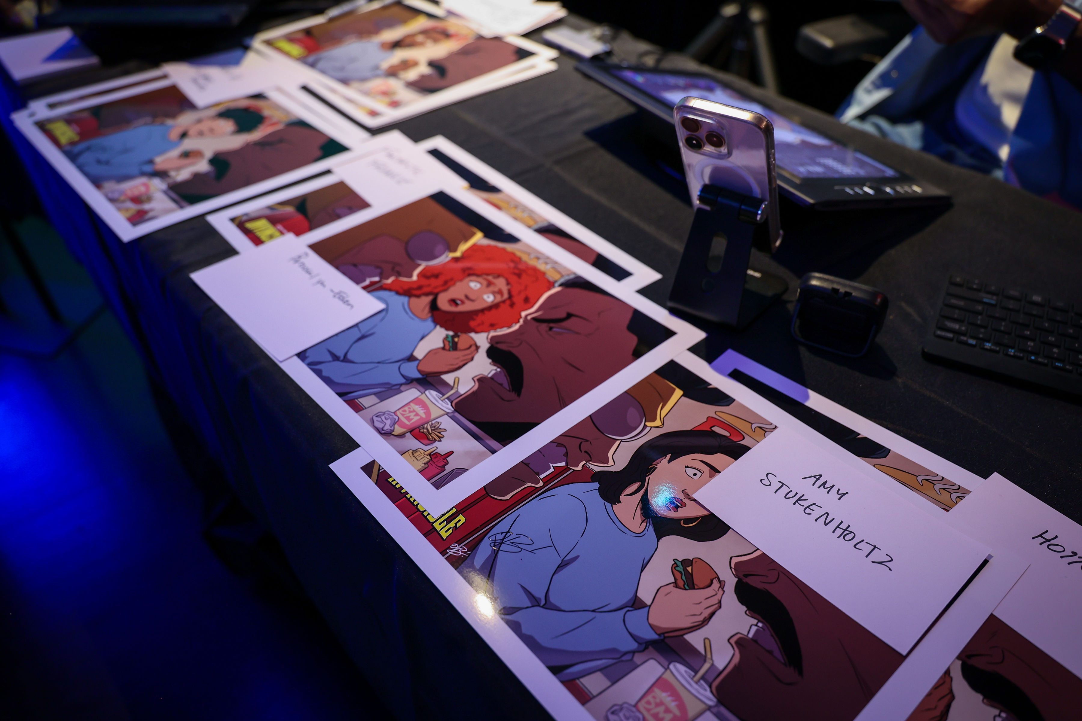 Glossy printed Invincible Season 4 illustrations laid out on a black tablecloth with handwritten guest name cards, a smartphone on a stand, and cool blue event lighting.