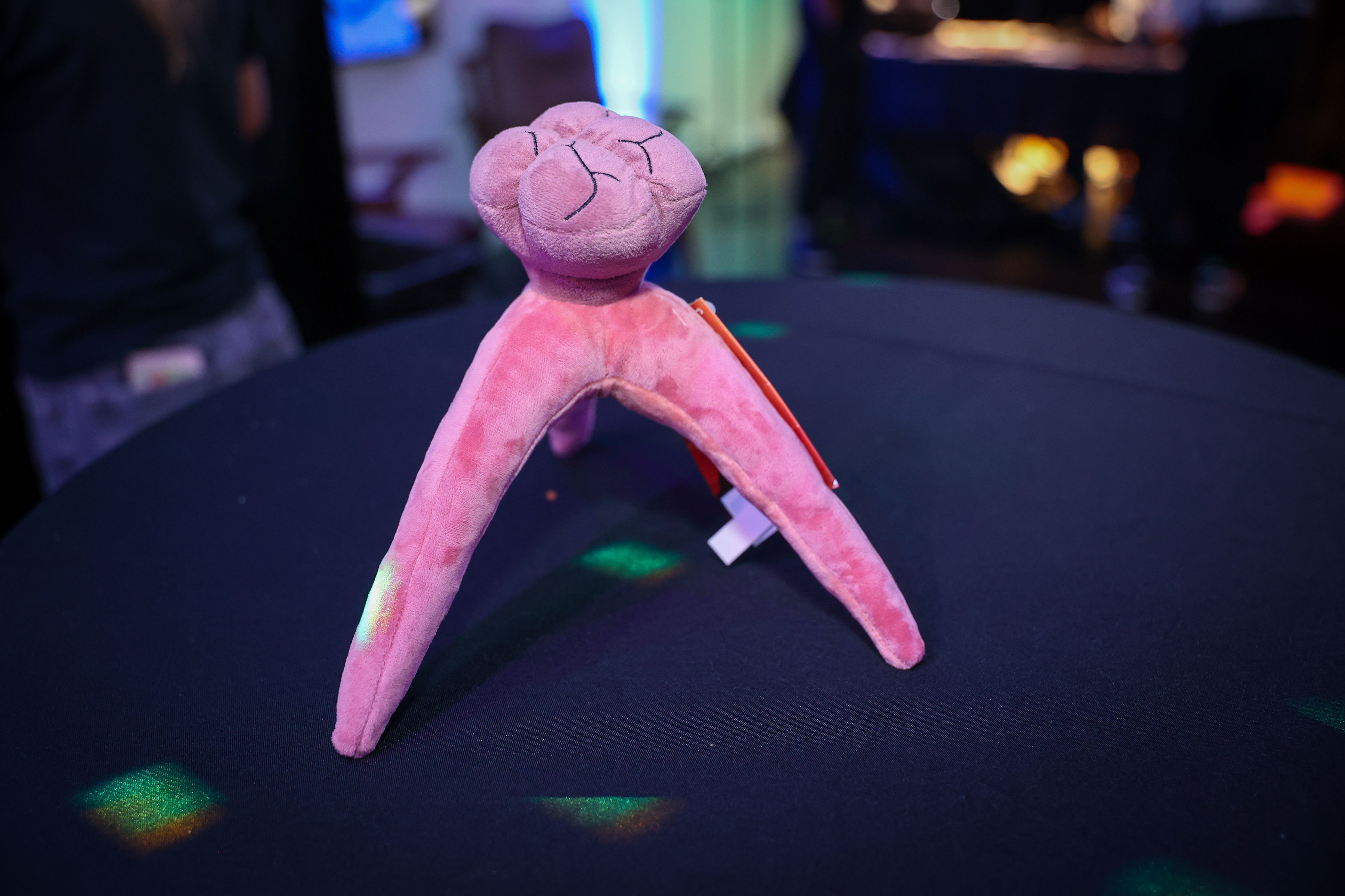 Pink, three-legged plush prop with visible stitched details standing on a black round cocktail table under colored event lighting and shallow depth of field.