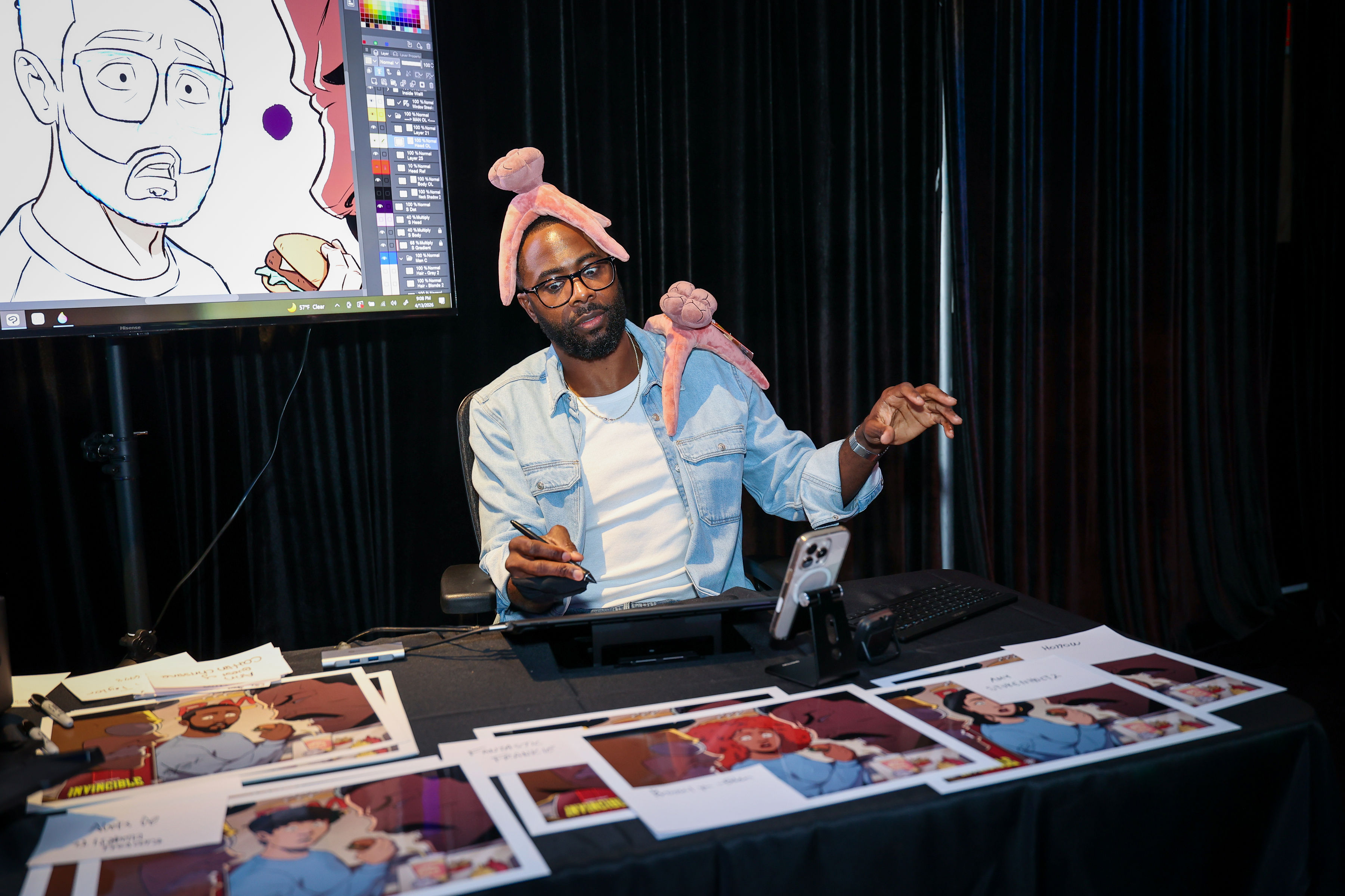 Seated illustrator working on a pen-display tablet with plush creatures on their head and shoulder, printed character illustrations spread across a table, and a reference image on a monitor.