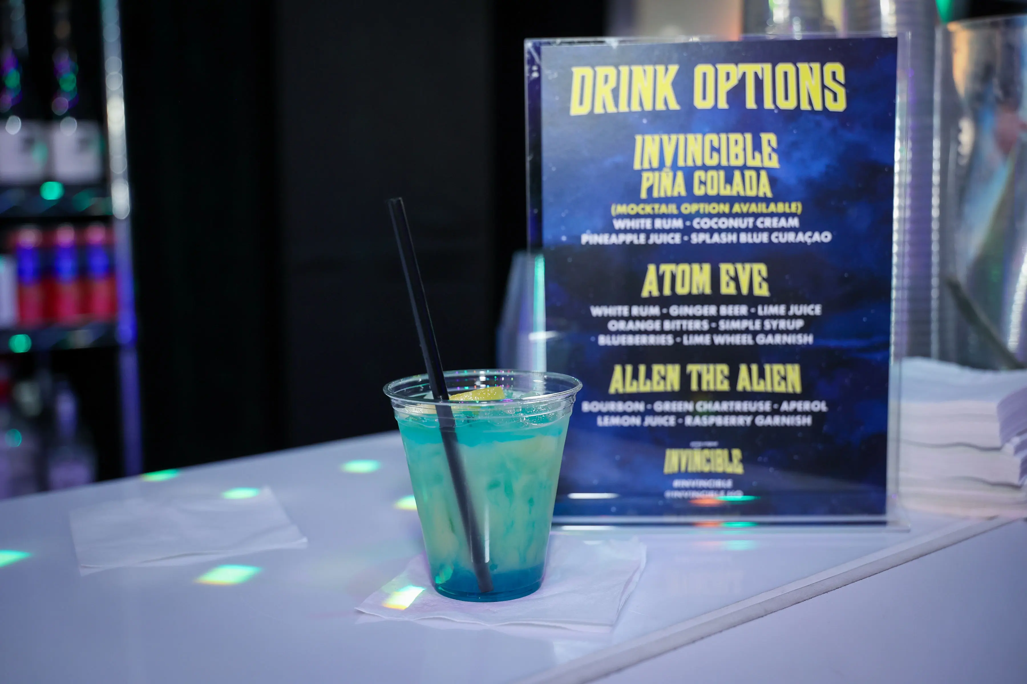 Blue-green layered cocktail in a clear plastic cup with a black straw on a white bar counter, next to a backlit yellow-on-blue 'Drink Options' menu listing Invincible Pina Colada, Atom Eve, and Allen the Alien.
