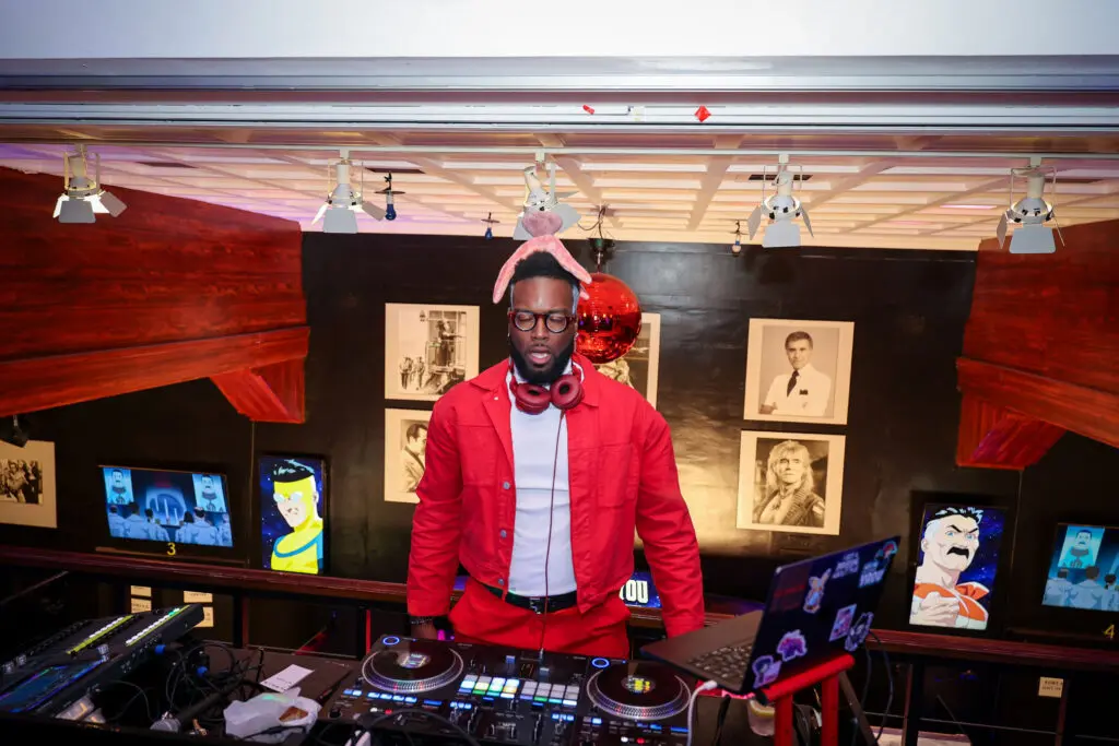 DJ wearing a red jacket and pink plush headgear stands behind a turntable in a mezzanine booth lit by gallery spotlights, with animated character screens and framed portraits on the black wall behind them.