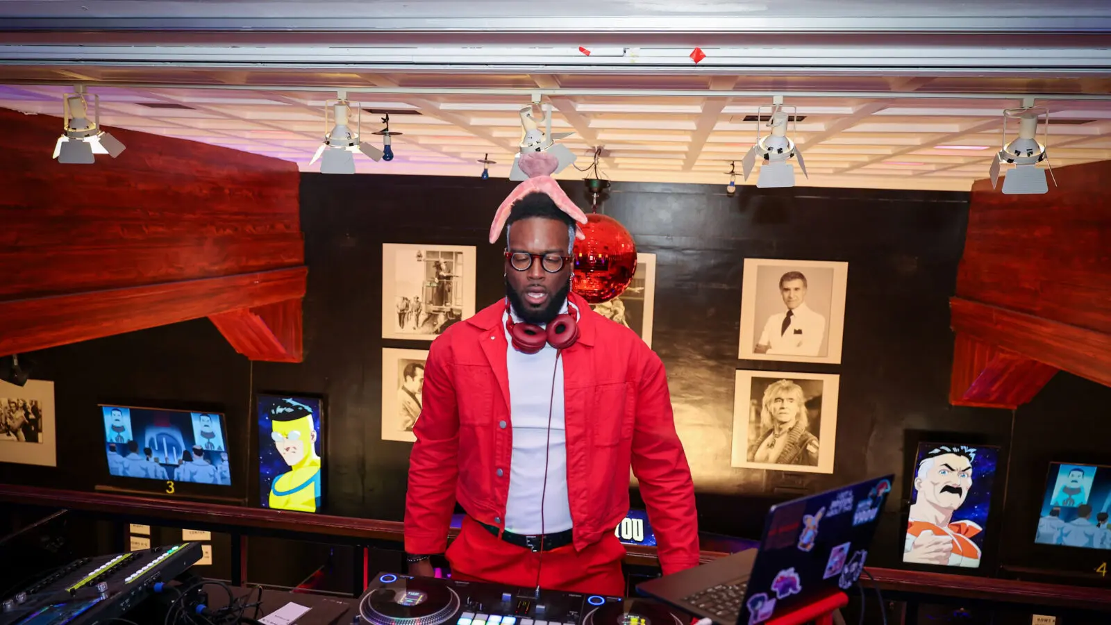 DJ wearing a red jacket and pink rabbit ears stands behind turntables and a sticker-covered laptop under a red disco ball, with framed portraits and animated character screens on the walls.