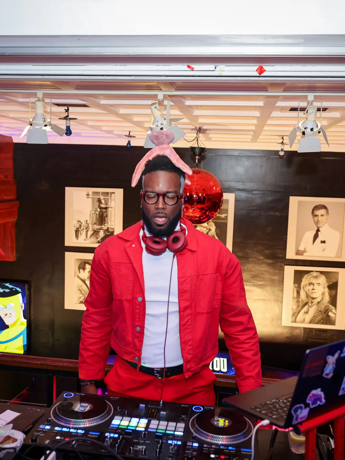 DJ wearing a red jacket and pink rabbit ears stands behind turntables and a sticker-covered laptop under a red disco ball, with framed portraits and animated character screens on the walls.