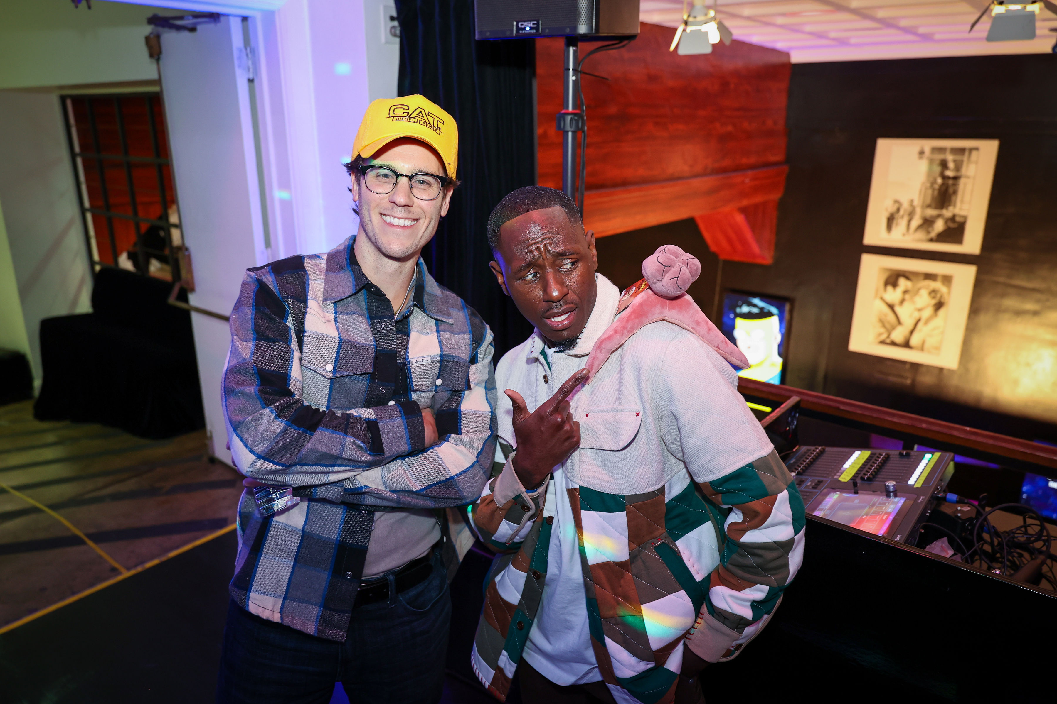 Two guests pose at a creator screening, one wearing a yellow CAT cap and glasses while the other points to a pink plush draped over his shoulder near a DJ mixing console.