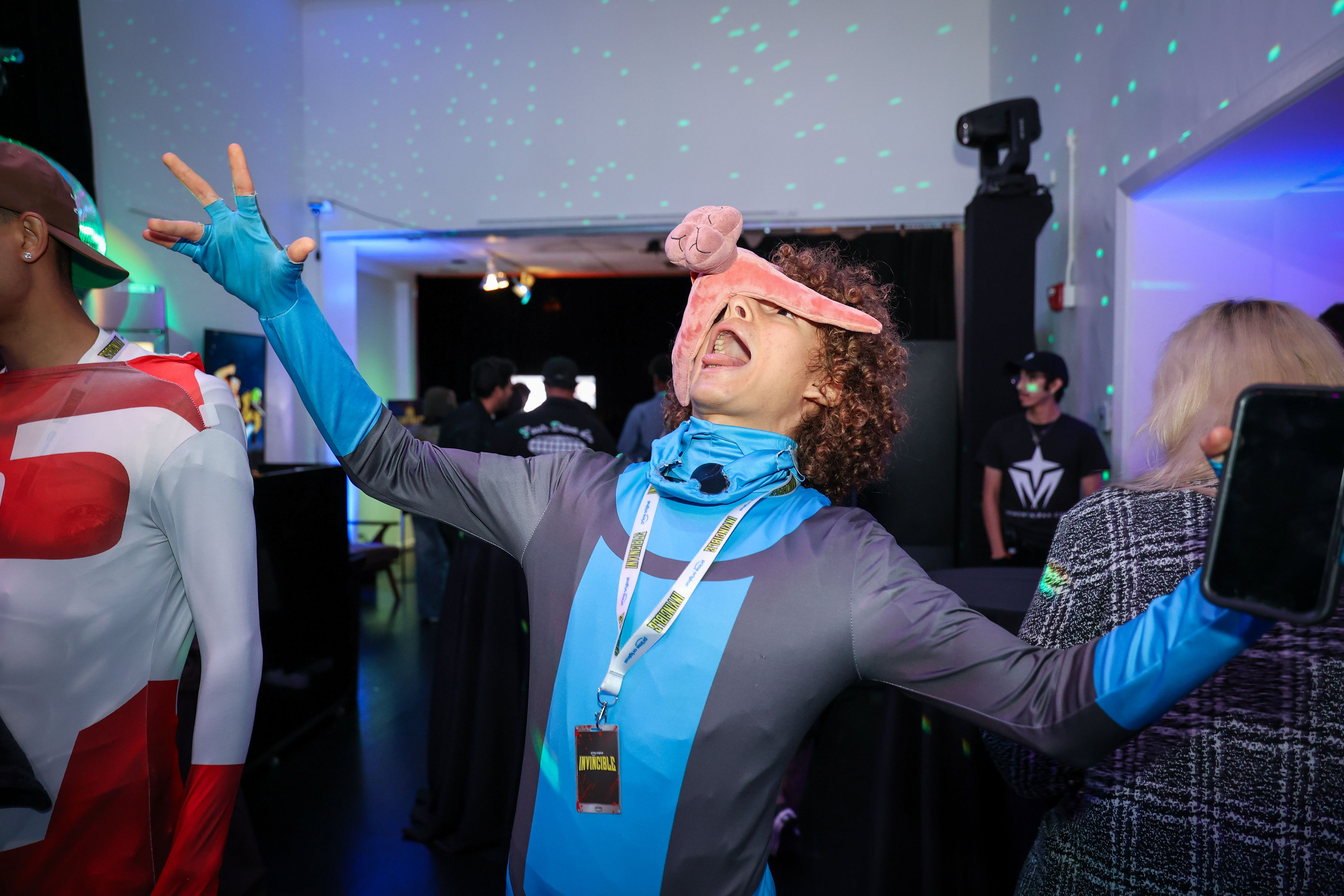 Person in a blue-and-gray superhero costume wearing a pink prosthetic across their face throws arms wide in a celebratory pose beneath green dot lighting while an Invincible badge hangs from their lanyard.