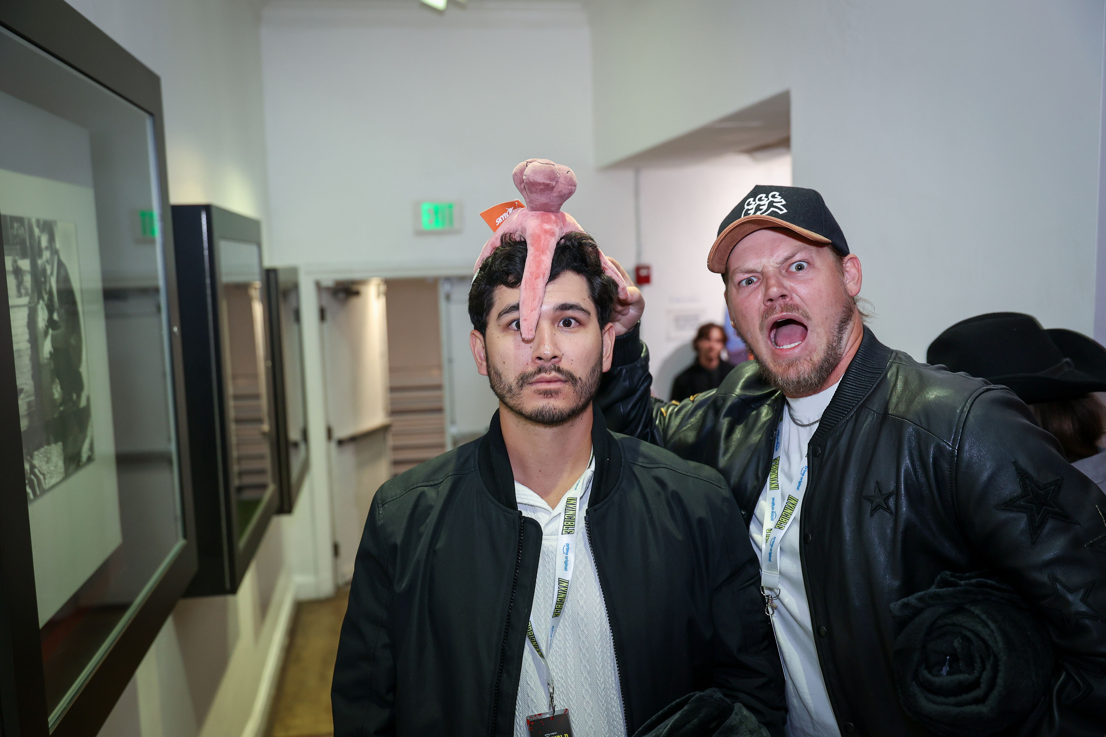 Two attendees in dark jackets stand in a hallway; one wears a pink plush creature on his head while the other makes an exaggerated shouting face beside him, shot for event documentation.