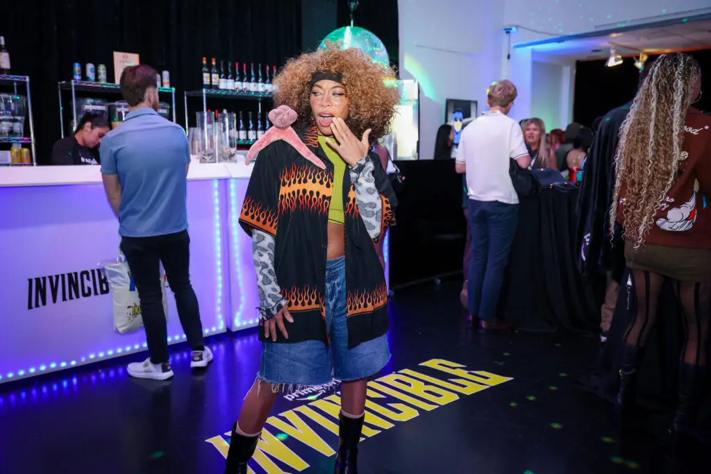 Person with voluminous curly hair wearing a flame-patterned jacket, denim shorts, and boots poses on a black floor decal reading INVINCIBLE, in a purple-lit event space with a backlit bar and disco ball.