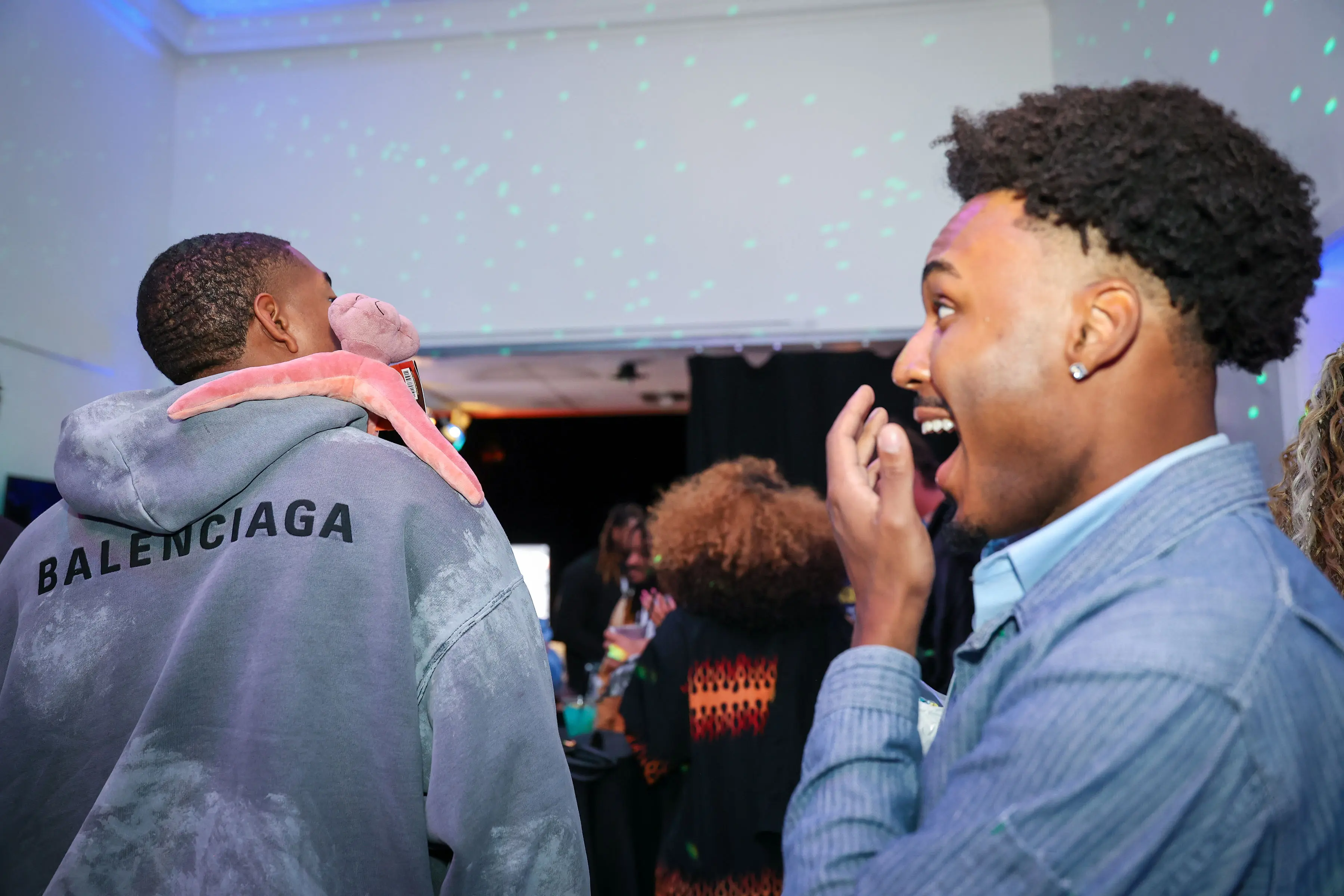 Two young men at an indoor event, one wearing a Balenciaga hoodie with a plush toy draped around his neck and another reacting with surprise under scattered green dot lighting.