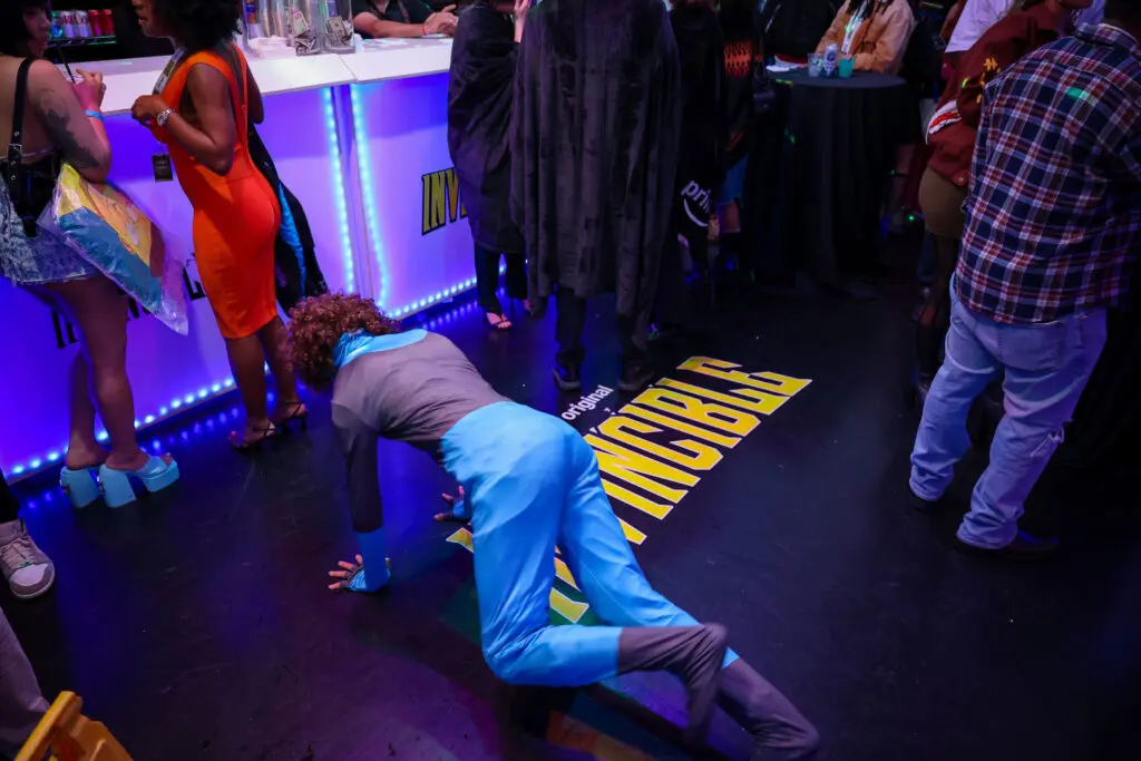 Person in a blue-and-black superhero costume crawling over a yellow INVINCIBLE floor decal while attendees gather at a lit concession bar with purple LED accents.
