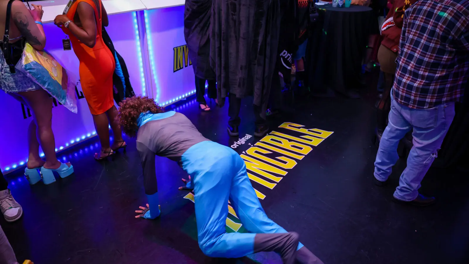 Cosplayer in a bright blue superhero costume crawling near a yellow INVINCIBLE floor logo, surrounded by attendees at a dim, blue-lit event bar.