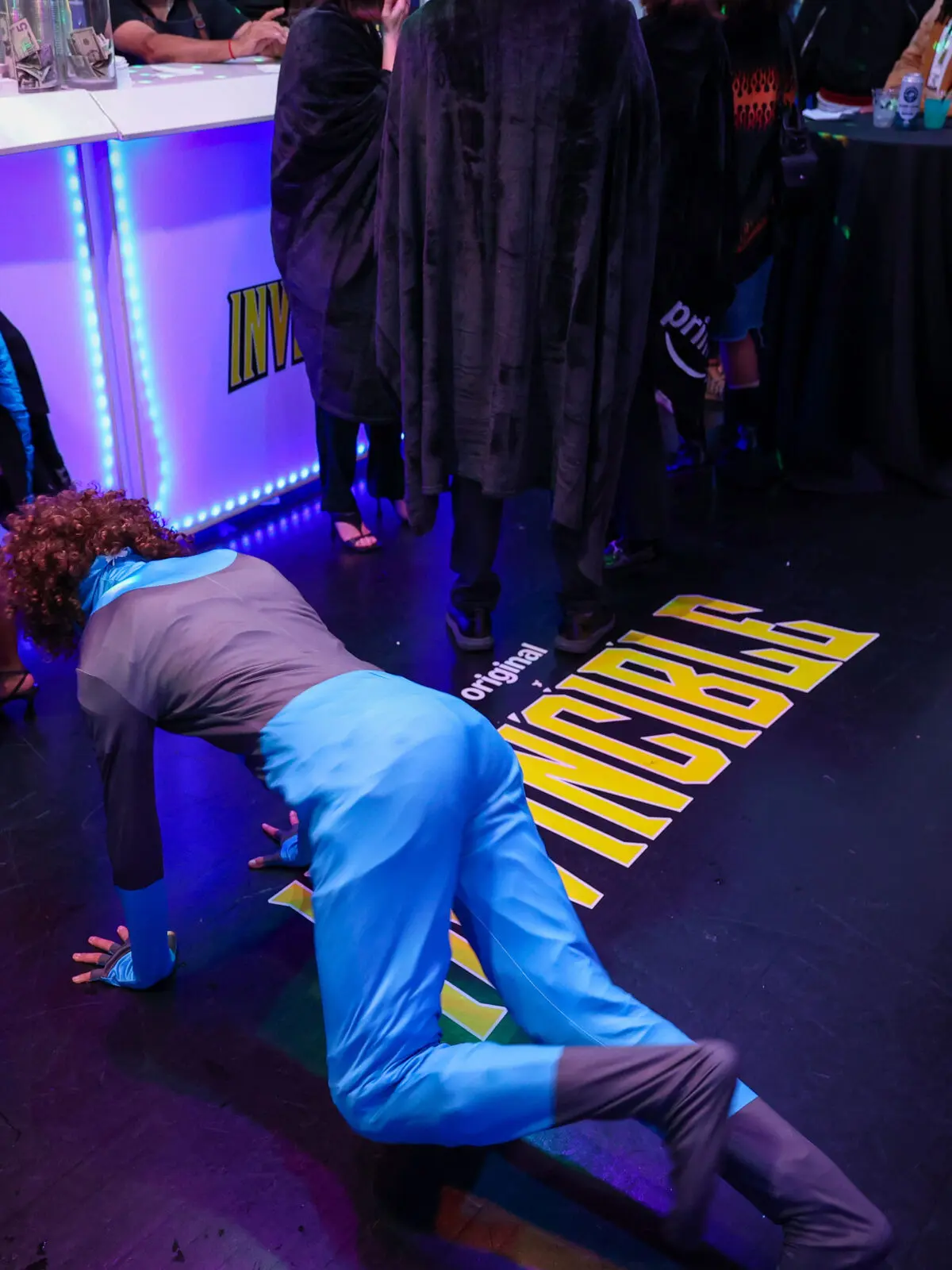 Cosplayer in a bright blue superhero costume crawling near a yellow INVINCIBLE floor logo, surrounded by attendees at a dim, blue-lit event bar.
