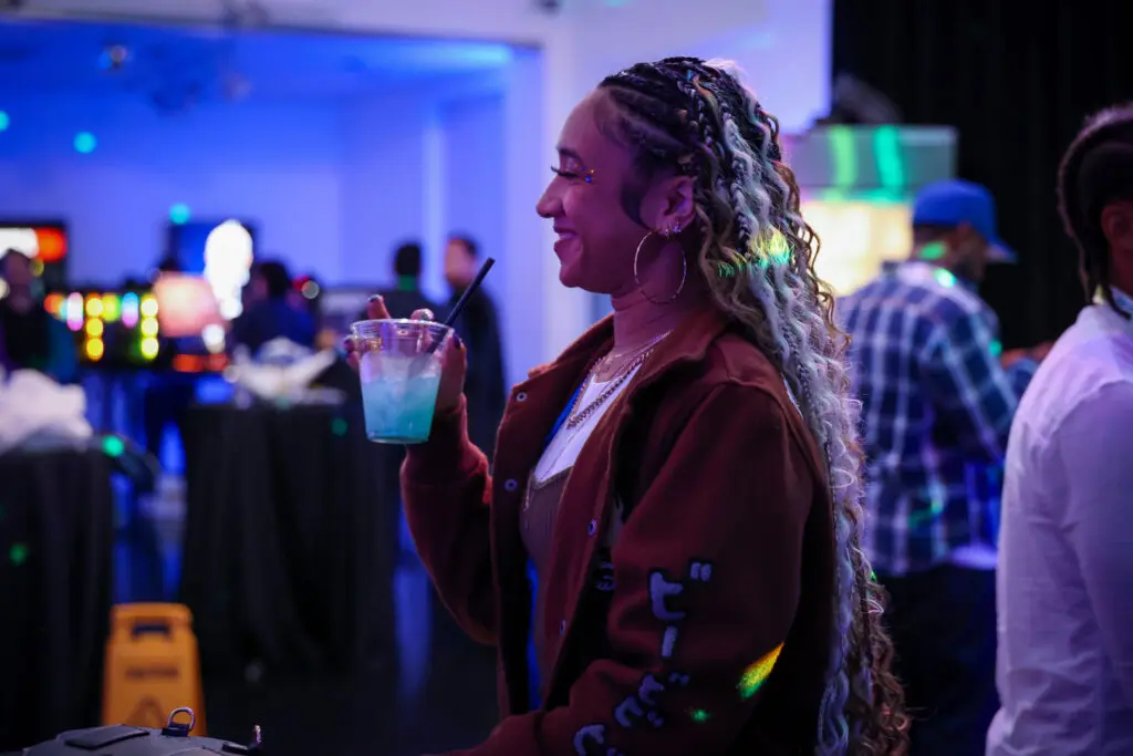 @jcheyenne_ smiles with a drink while attending the Invincible Season 4 creator screening, surrounded by other guests and colorful event lighting.