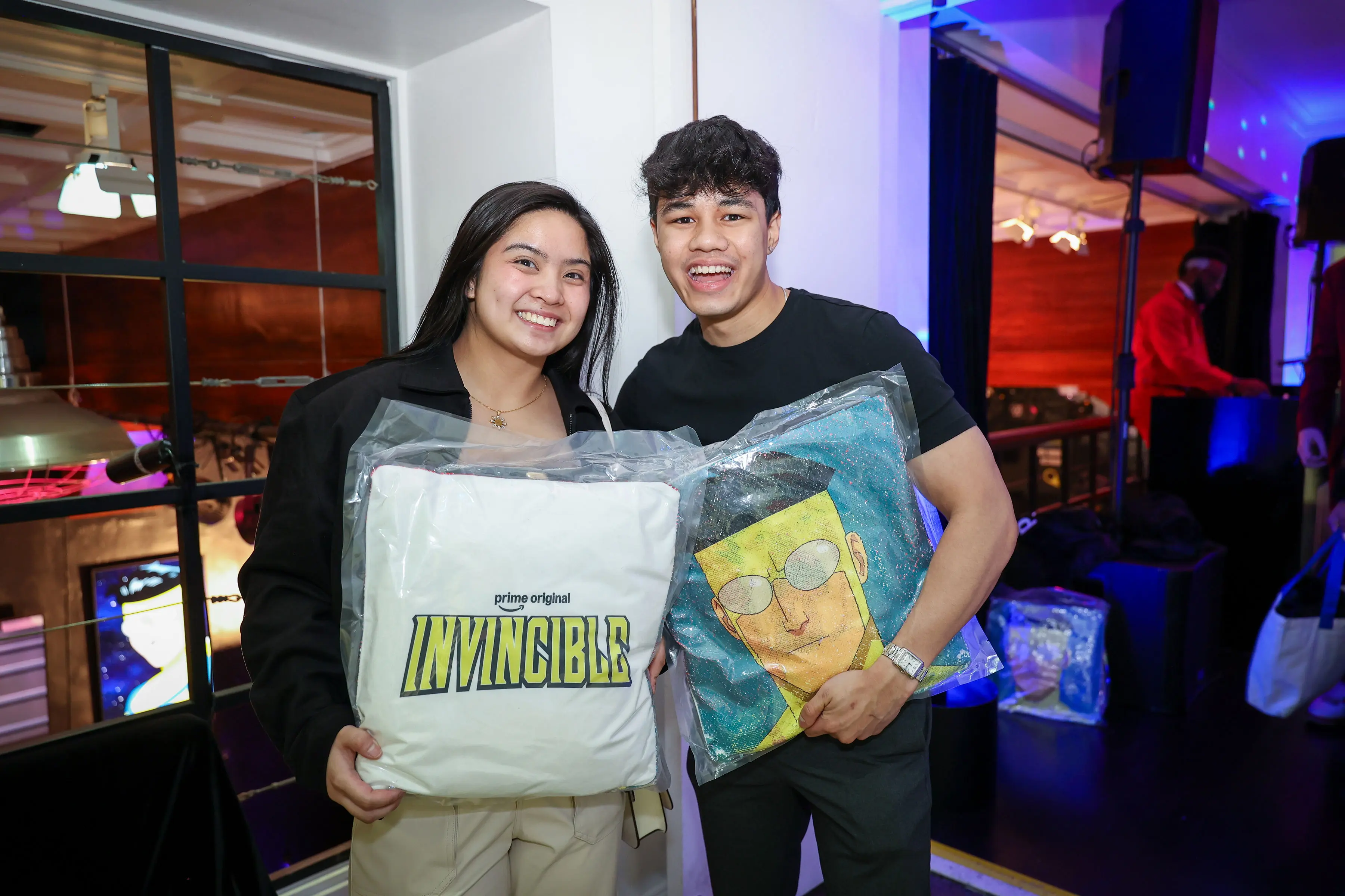 Two attendees at the Invincible creator screening hold branded pillows, one featuring the Invincible wordmark and one featuring a comic-style character illustration.