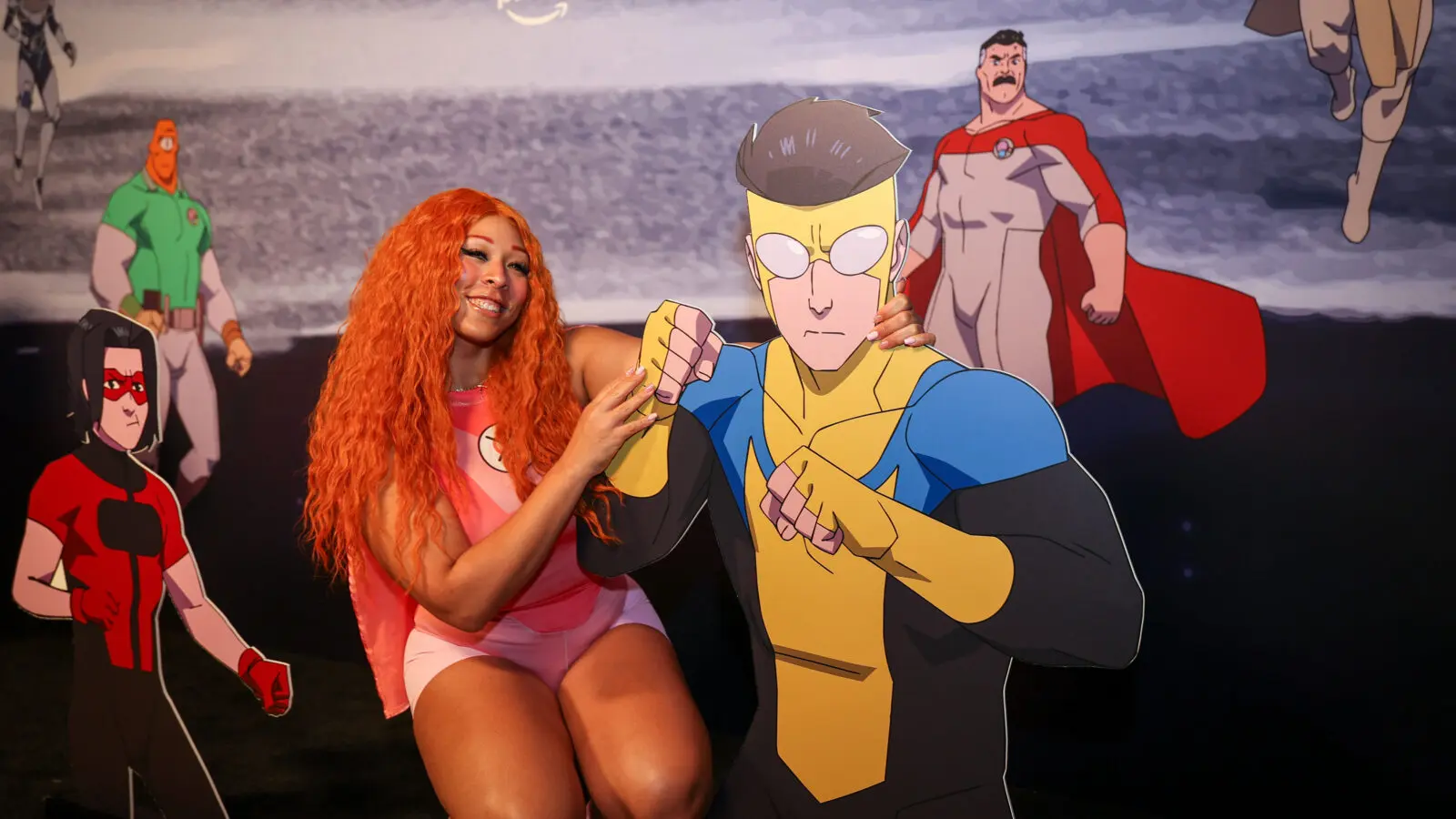 Smiling woman with long orange hair crouches beside a life-size cardboard cutout of Invincible in a yellow-and-blue suit, posed against a branded backdrop featuring other Guardians of the Globe characters.