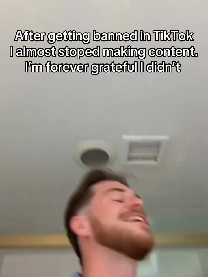 Man with red beard laughing in a vertical selfie frame, ceiling vents visible above him and bold black text overlay reading 'After getting banned in TikTok I almost stoped making content. I'm forever grateful I didn't.'