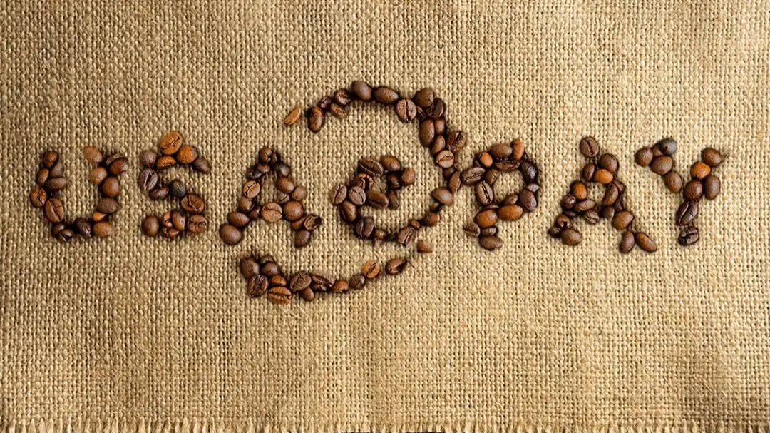 Coffee beans arranged to spell 'USAEpay' on a rectangular burlap strip, with loose beans above and a yellow cup rim and spoon visible along the lower edge.
