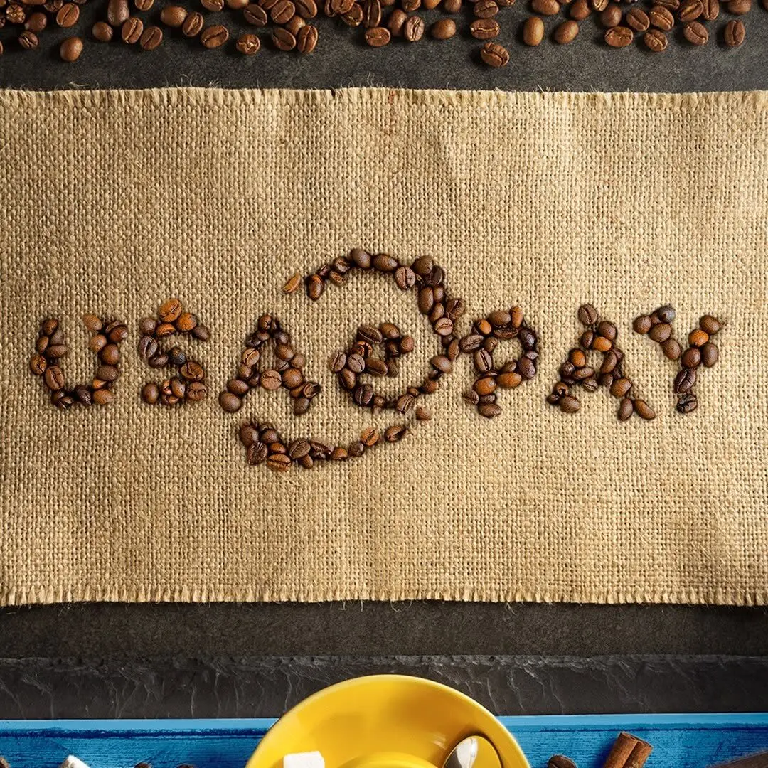 Coffee beans arranged to spell 'USAEpay' on a rectangular burlap strip, with loose beans above and a yellow cup rim and spoon visible along the lower edge.