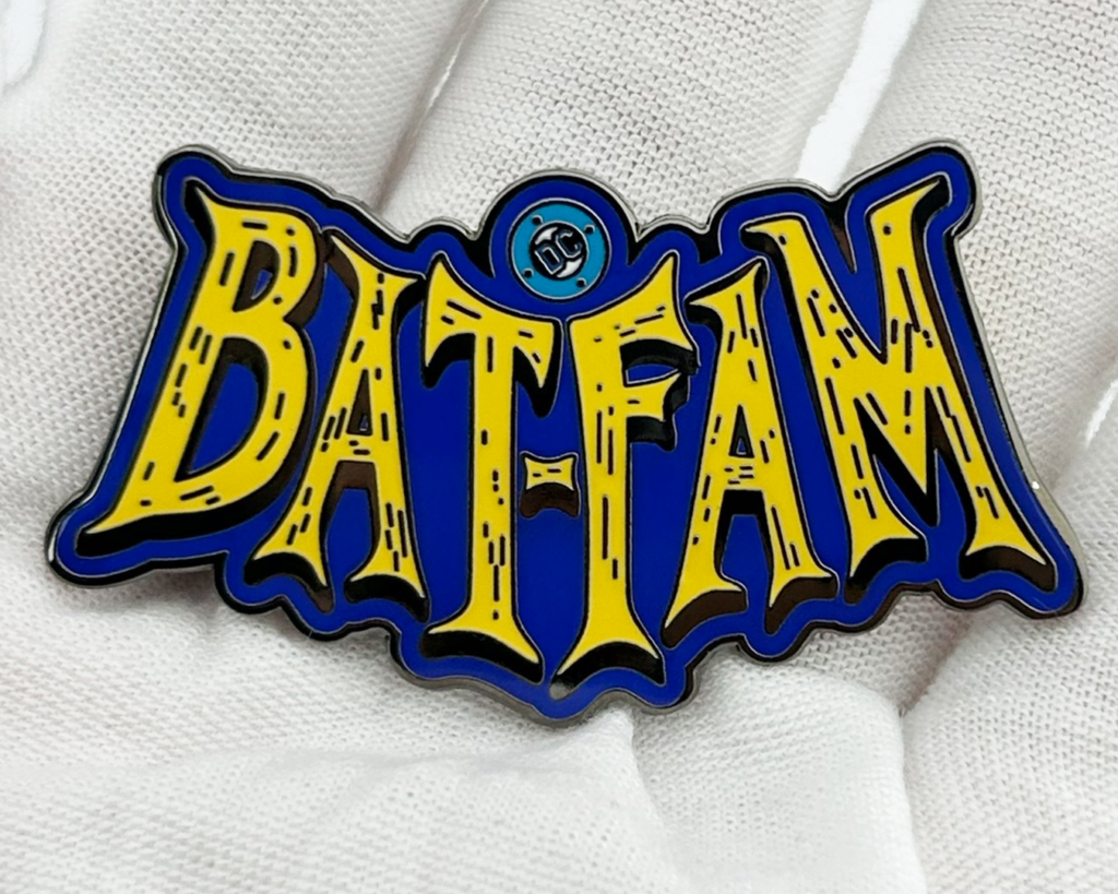 Glossy enamel lapel pin reading “BAT-FAM” in bright yellow lettering with a deep blue background and a small circular DC mark, photographed resting on white gloved fabric.