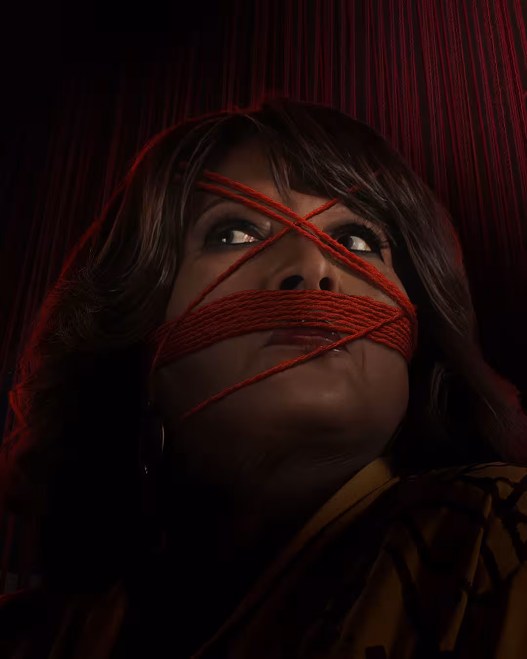 Low-angle close-up of Athena with red yarn wound across her mouth and eyes, vertical red threads in the background, dramatic shadowed lighting emphasizing texture and tension.