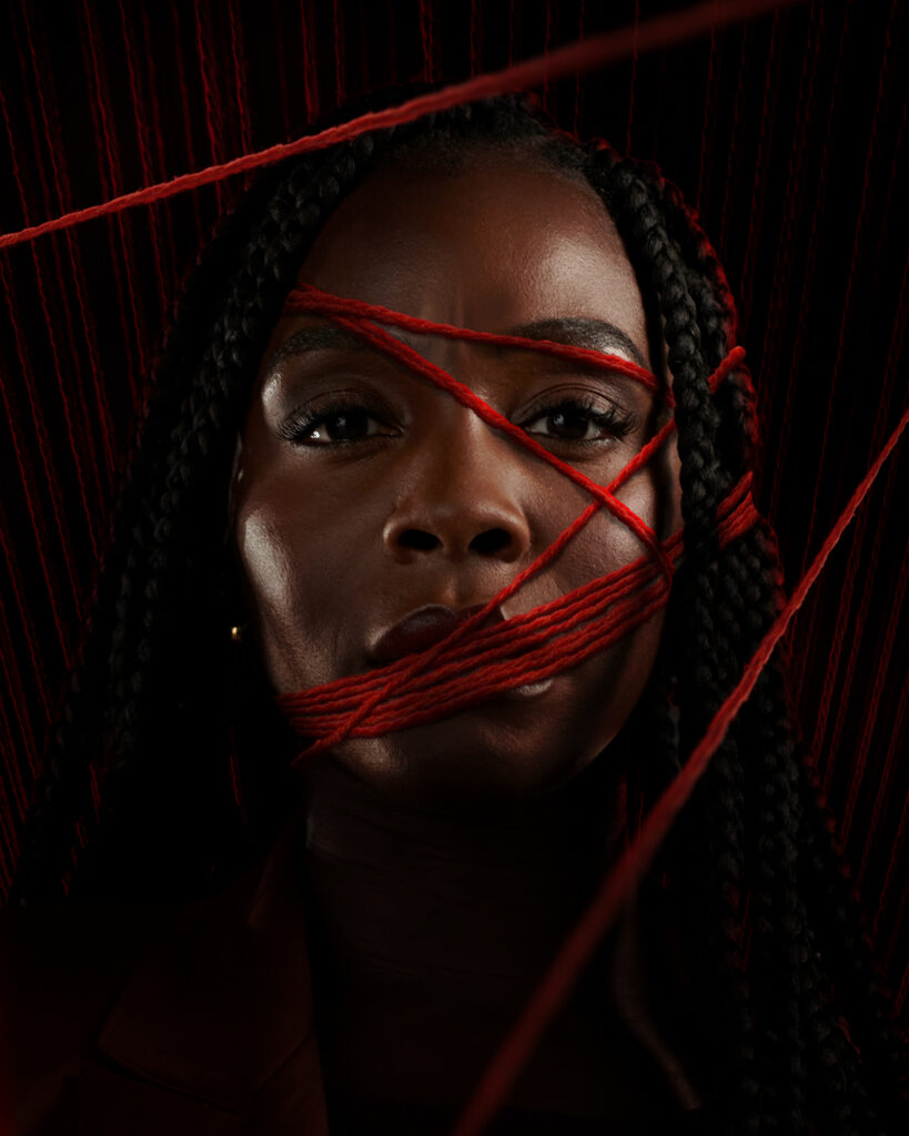 Close-up portrait of Deborah Ayorinde surrounded by strands of red yarn for Prime Video’s THEM: The Scare, set against a dark, textured background.