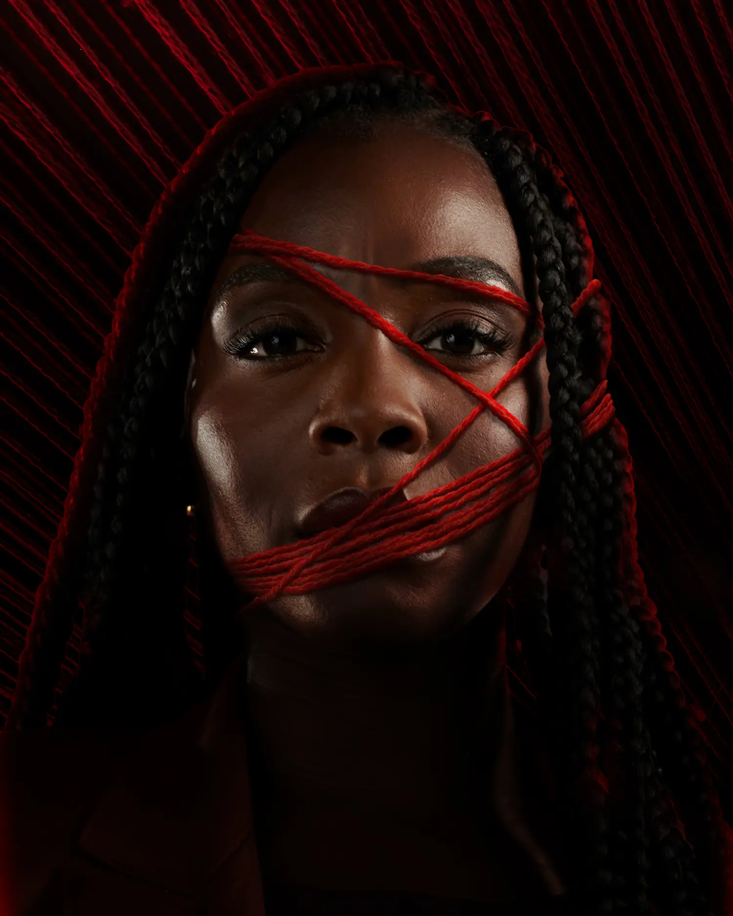 Close-up portrait of a Black woman with braided hair, red yarn wrapped tightly across her mouth and diagonally over her face, set against a dark background with radiating red threads.