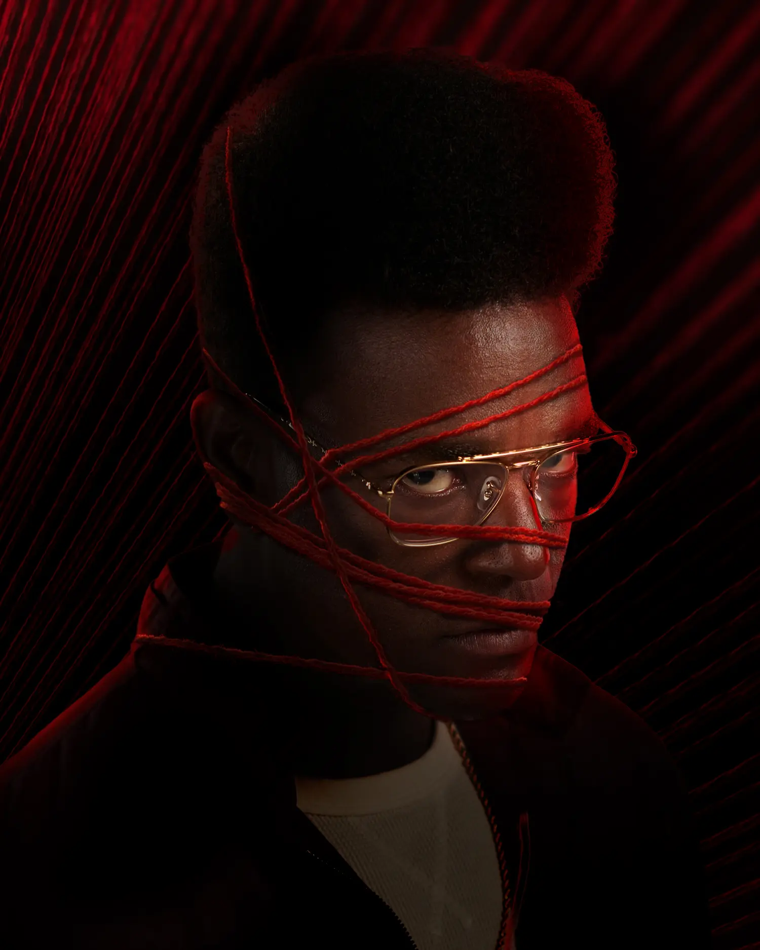 Close-up portrait of Edmund Gaines with red yarn tightly wrapped across his face and gold-rimmed glasses, set against a deep red threaded background that echoes tangled, frayed fabric.