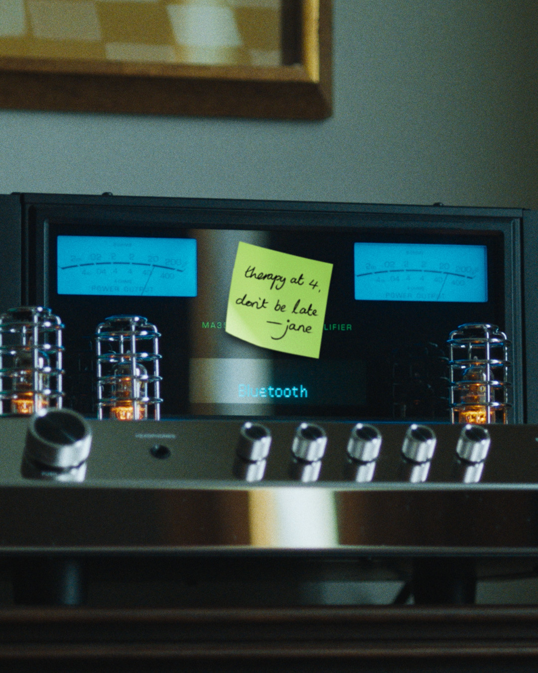 Front-facing hi-fi amplifier with glowing vacuum tubes, two blue VU meters, a digital 'Bluetooth' display, and a yellow sticky note reading 'therapy at 4, don't be late — jane' above the controls.