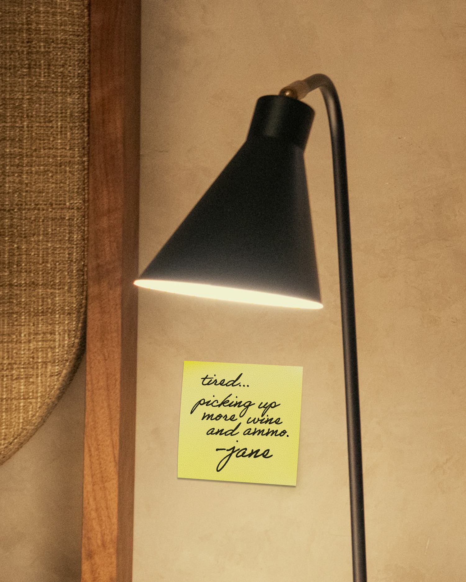 Floor lamp casting warm light on a beige wall with a yellow handwritten sticky note reading "tired... picking up more wine and ammo. -jane" and woven upholstery at left.