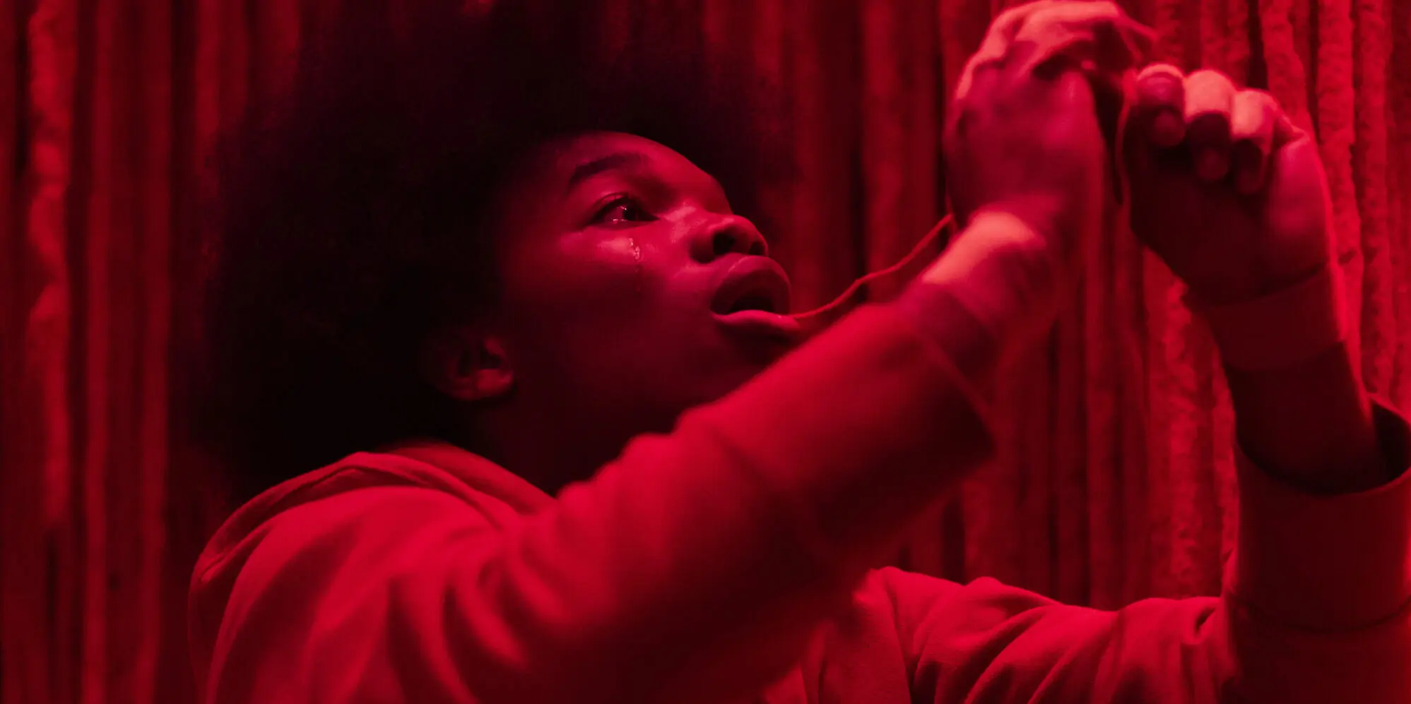 Young Black person bathed in intense red light, looking upward with a single tear while pulling a strand of yarn or thread against a textured vertical fabric backdrop.