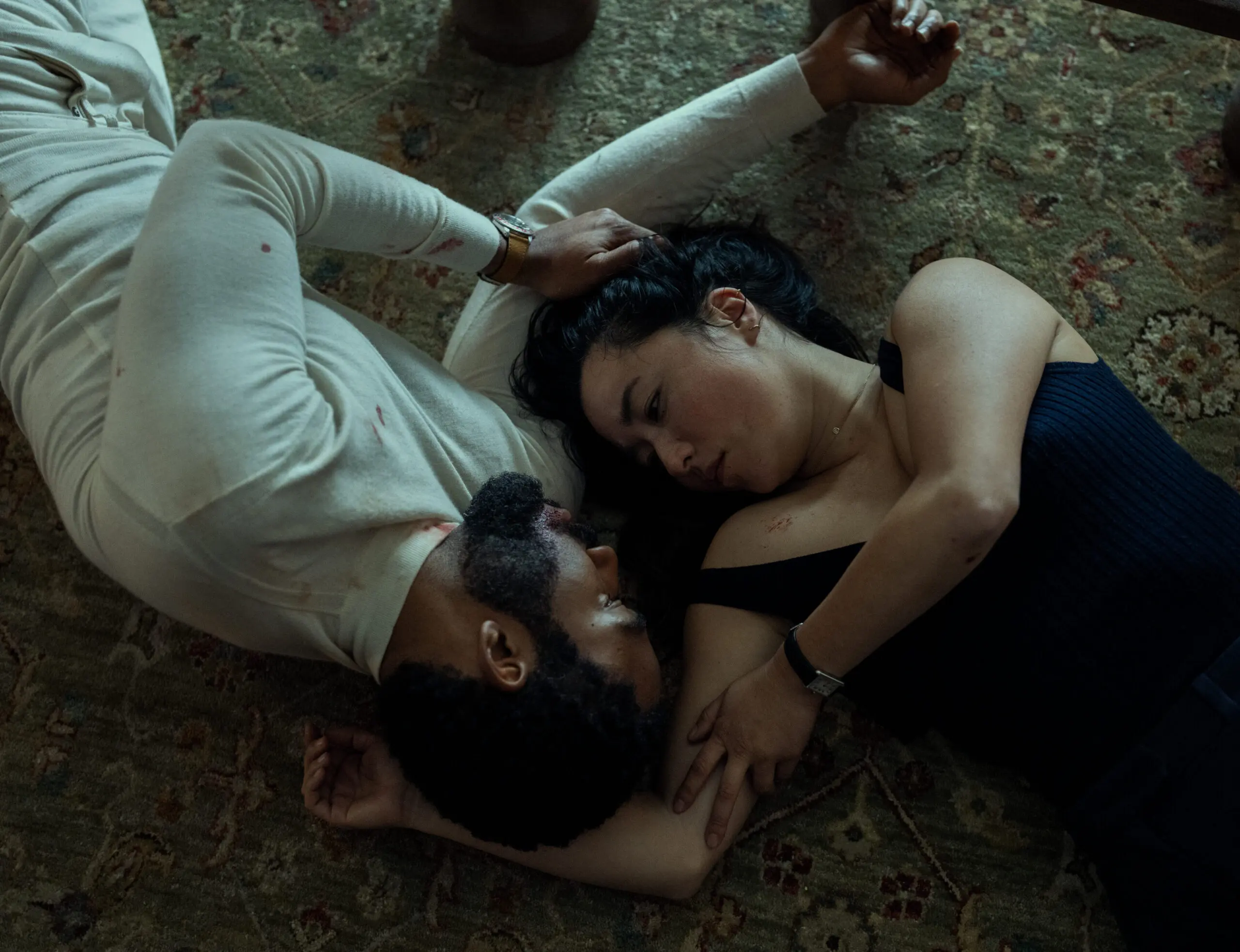 John and Jane Smith lie face-to-face on a patterned rug, their bodies close with his white shirt faintly stained, framed in low, cinematic light that feels intimate and tense.