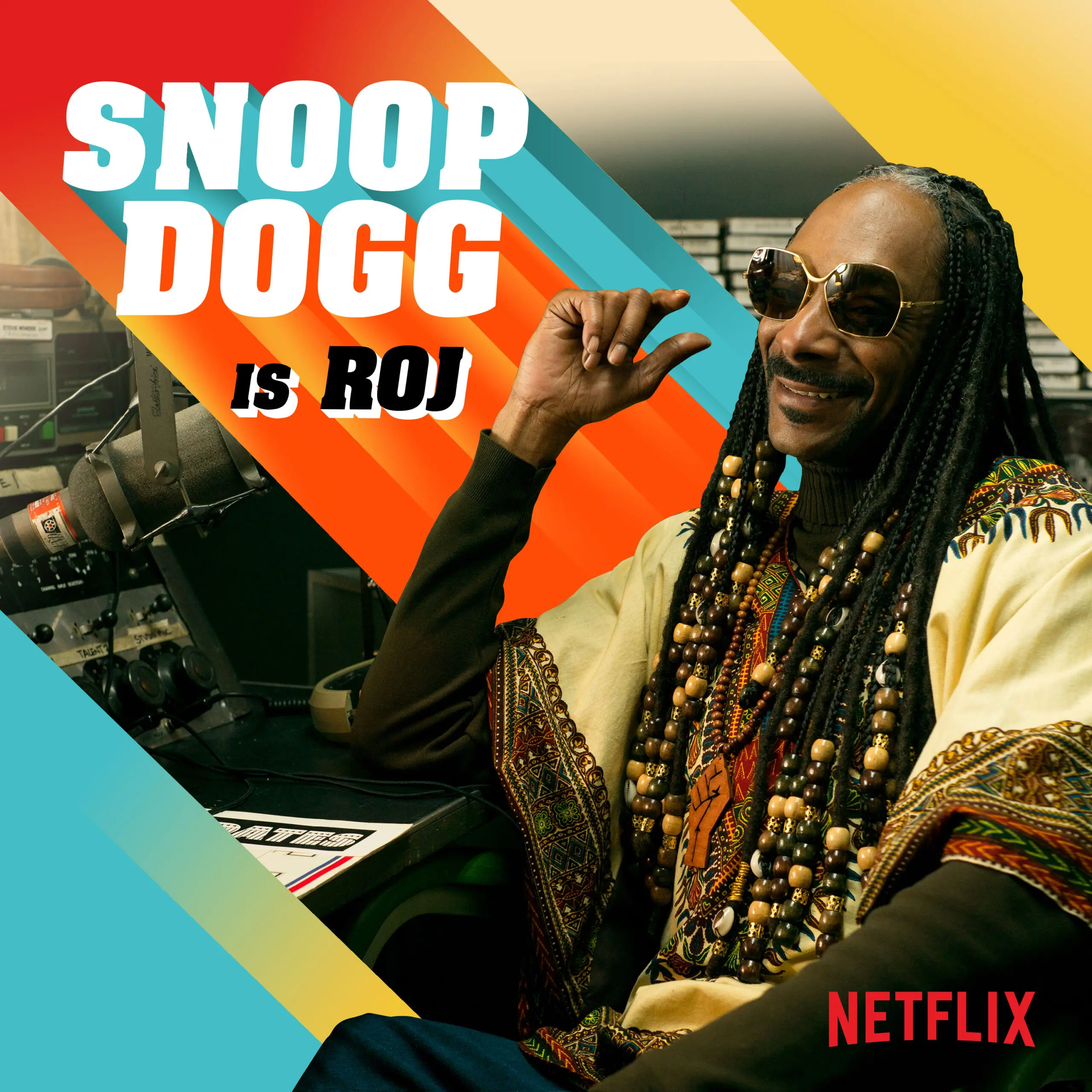 Actor in character as 'Roj' seated in a radio booth wearing beaded braids, sunglasses and a patterned dashiki, set against bold diagonal orange, teal and yellow 1970s–style stripes.