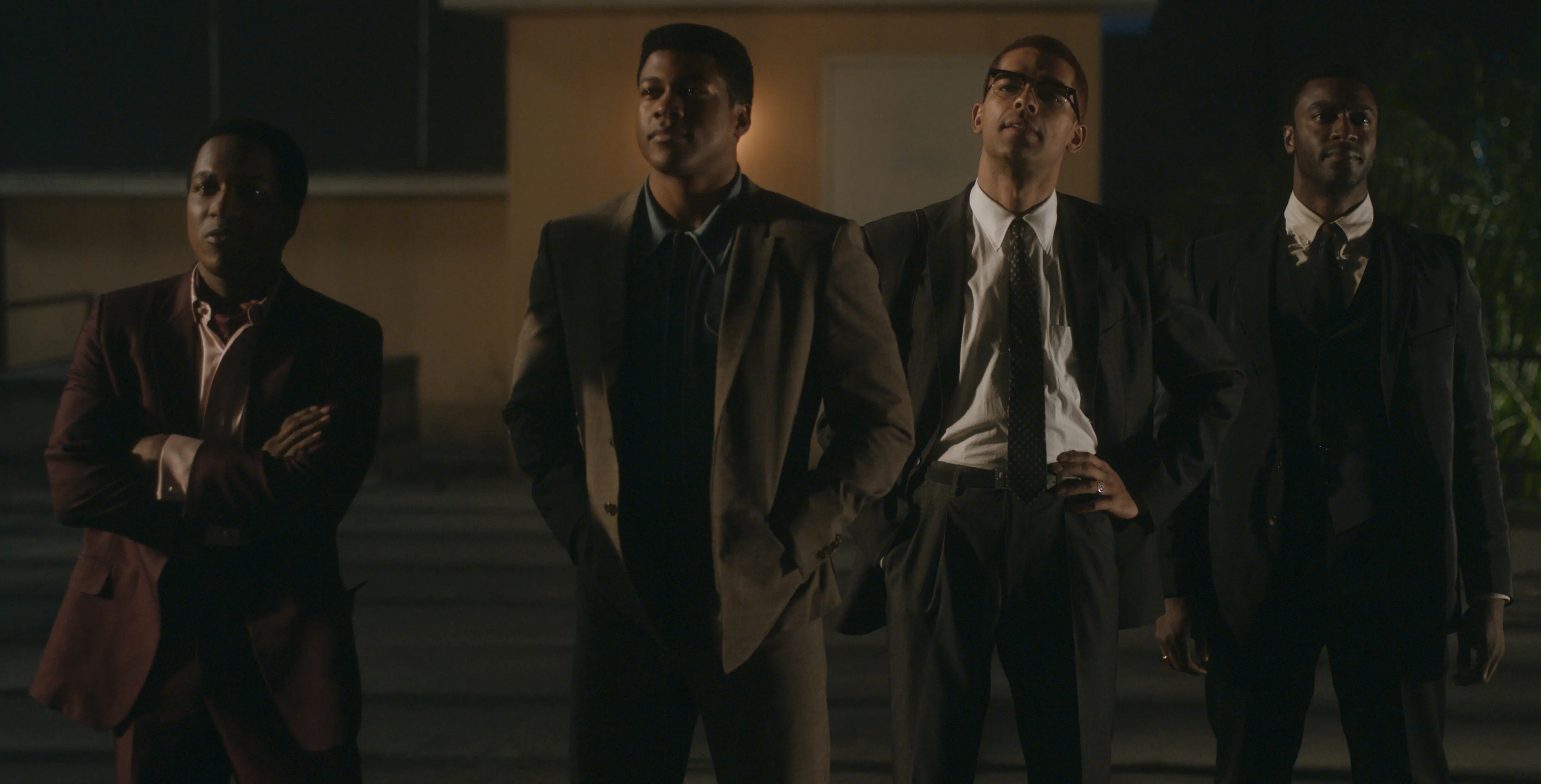 Four Black men in 1960s suits stand side-by-side outside a motel at night, lit by warm facade light and deep shadows that emphasize their silhouettes and posed stillness.