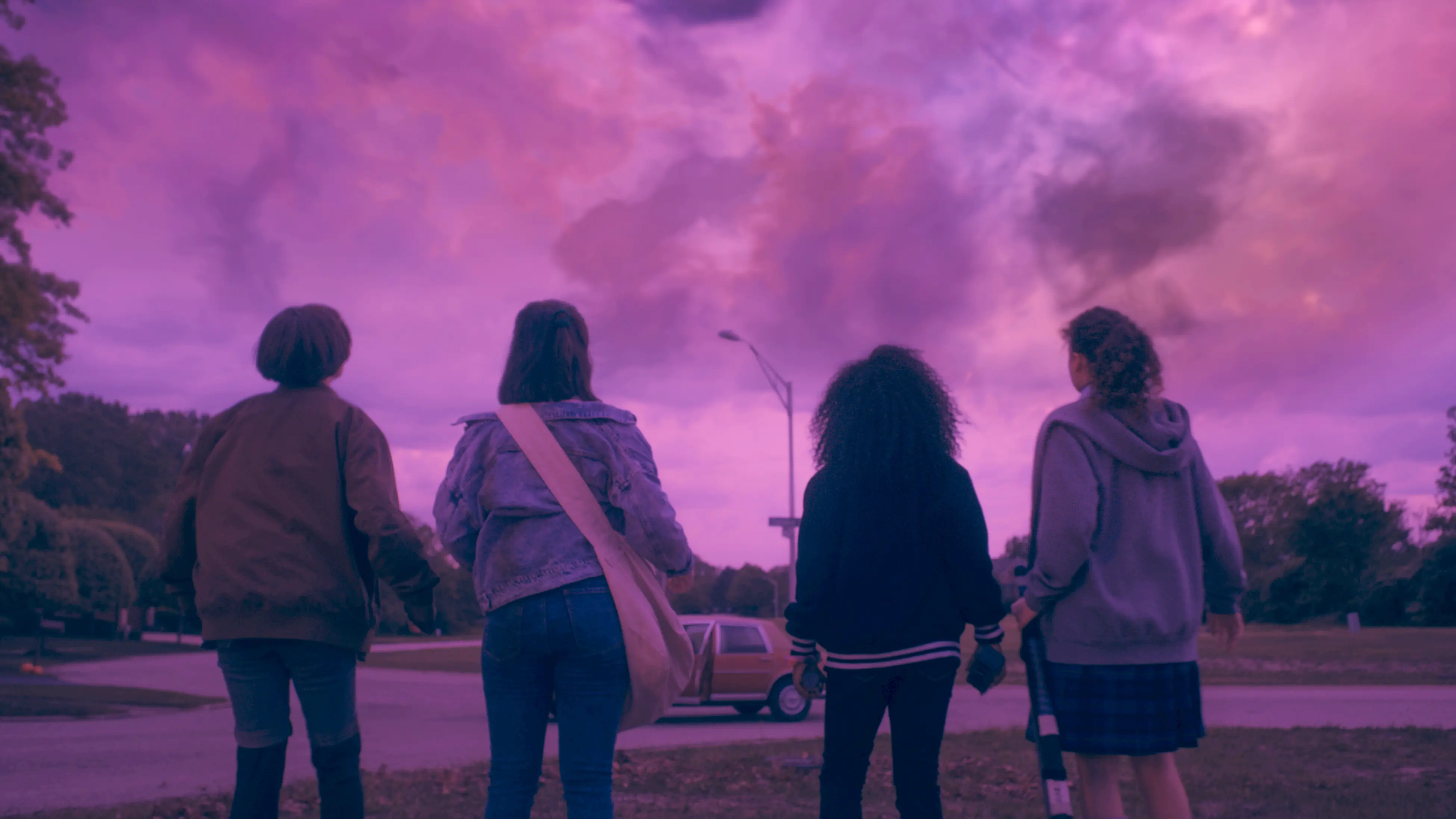 Four paper-delivery girls stand with their backs to camera on a quiet suburban street at dawn, silhouetted against a magenta-purple sky with a parked car and trees in the background.
