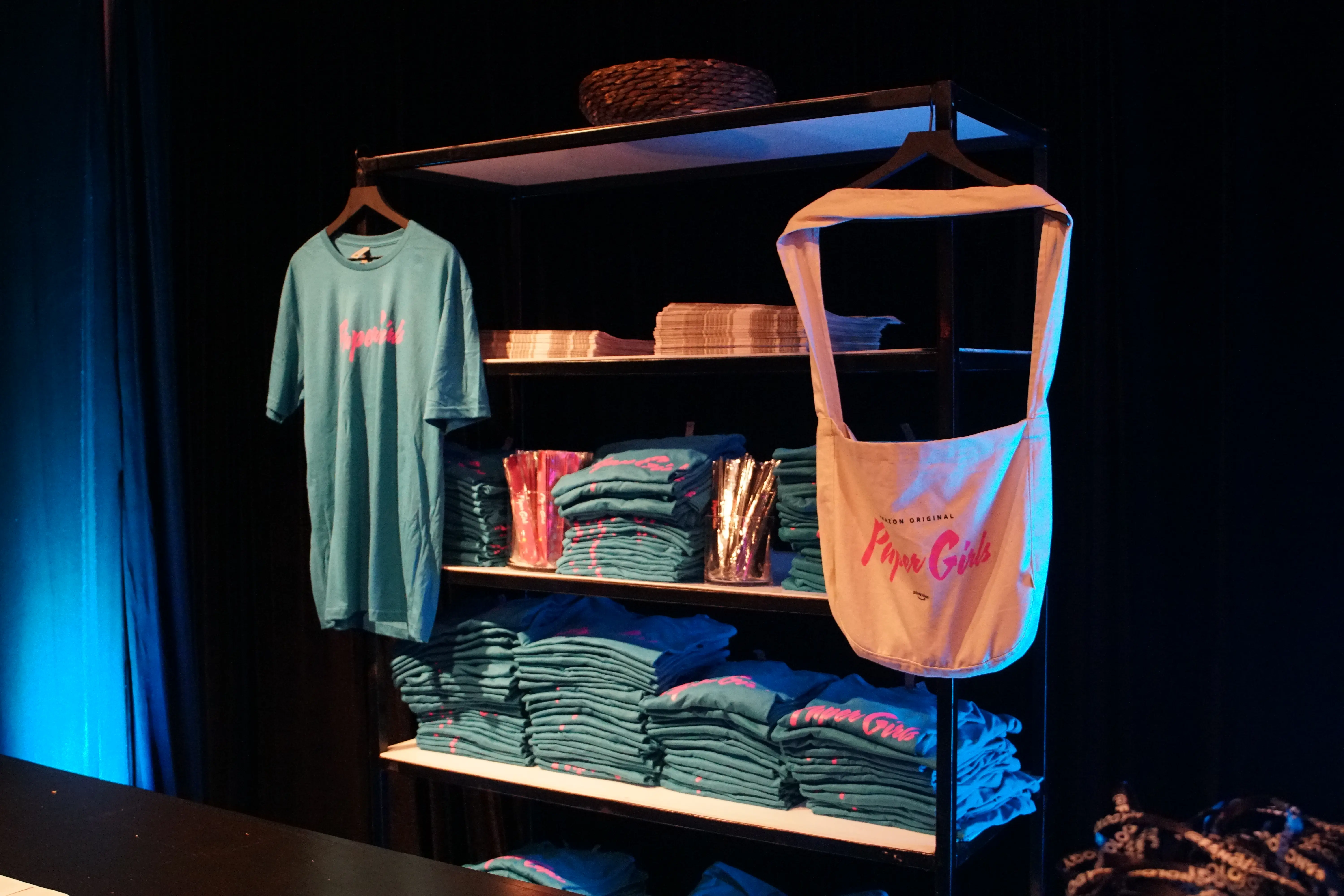 Turquoise Paper Girls t-shirts stacked on metal shelving with a hanging tee and a canvas tote printed in pink script, folded newspapers on a middle shelf under cool blue event lighting.