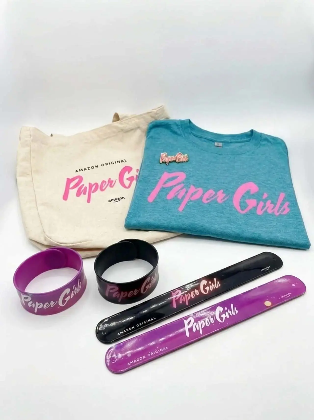 Teal folded T‑shirt with pink 'Paper Girls' script beside a cream canvas tote stamped 'Amazon Original' and 'Paper Girls', two silicone wristbands and two purple-and-black slap bracelets on white.