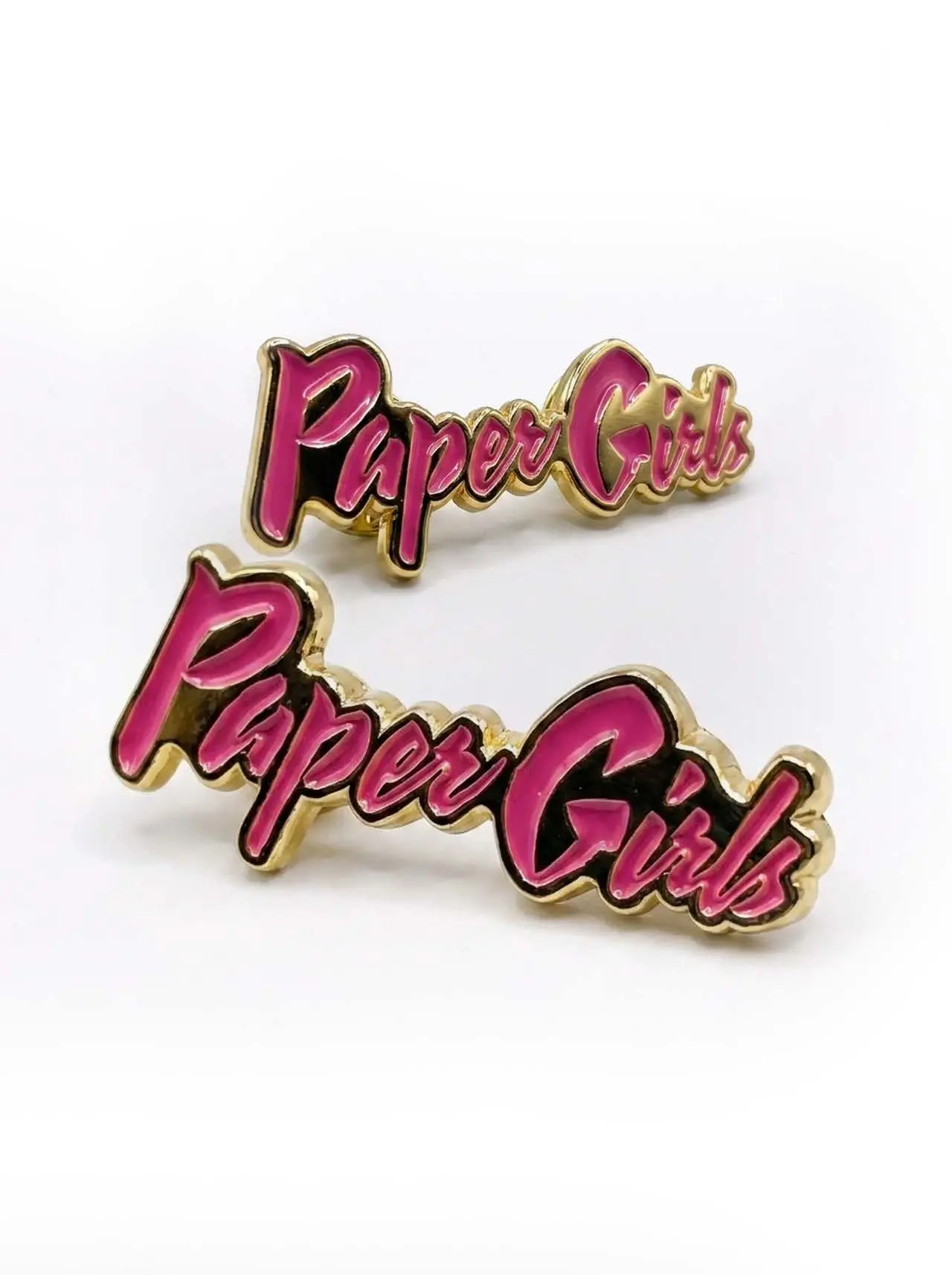 Two pink-script enamel lapel pins reading “Paper Girls” with gold metal edges on a white background, one pin in sharp focus and a second pin slightly blurred behind it.