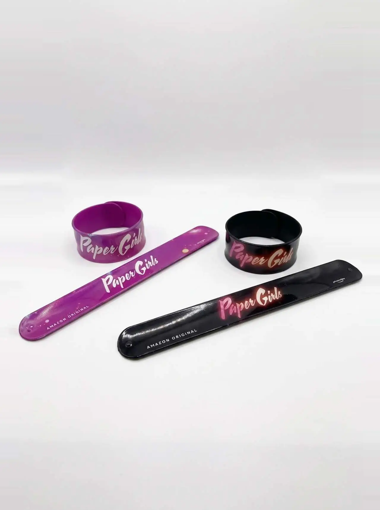 Four Paper Girls wristbands, two glossy slap bracelets and two looped cuff bands, laid on a white background in purple and black with a neon-pink Paper Girls script and small Amazon Original text.