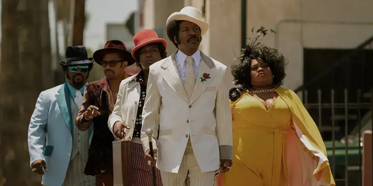 Rudy Ray Moore in a white suit and hat leads a flamboyantly dressed group—three men in colorful 1970s suits and a woman in a yellow dress—striding down a sunlit street with a cane and suitcase.