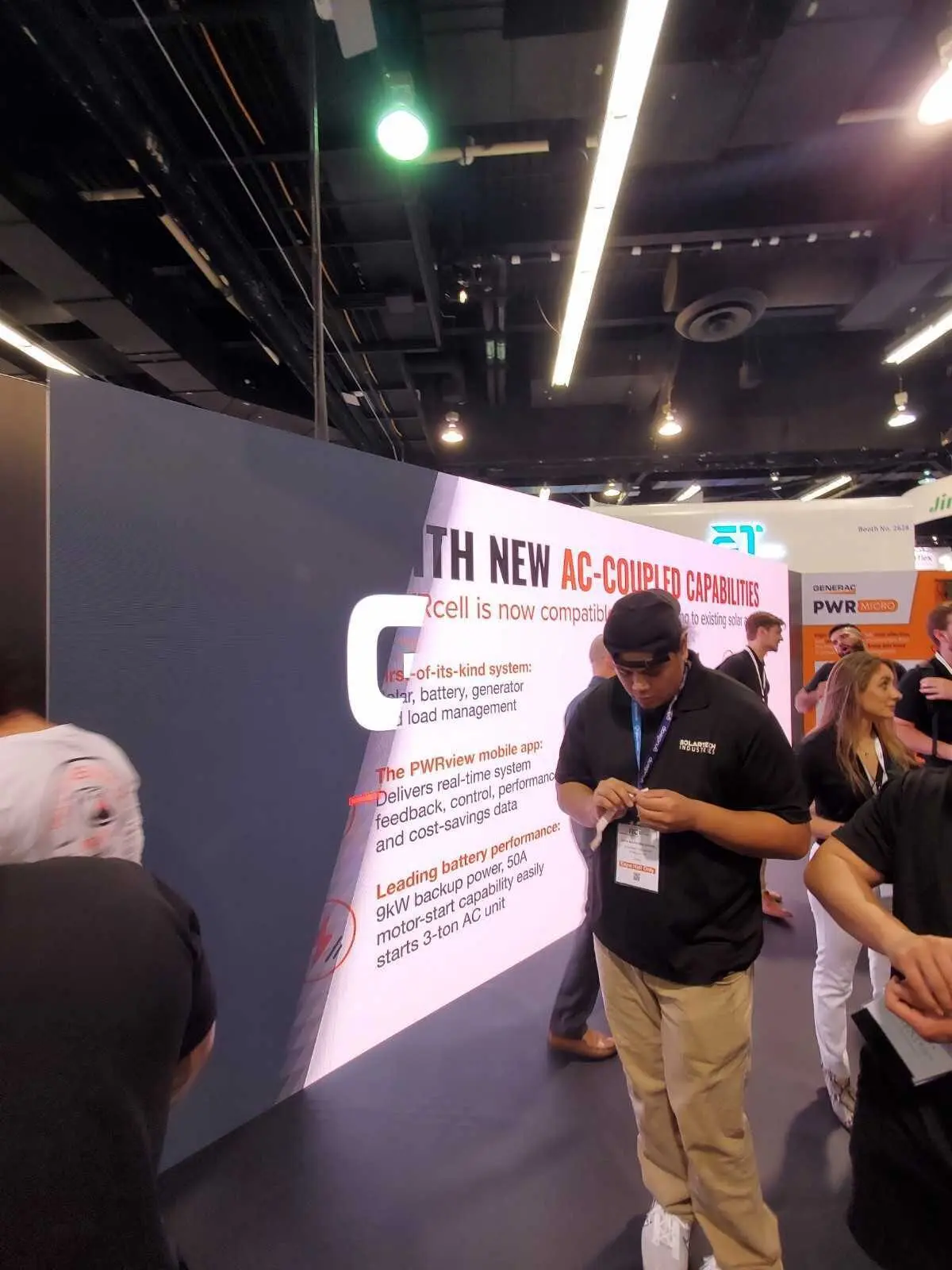 Curved LED video wall at a Generac trade-show booth displaying large copy about AC-coupled PWRcell capabilities, with attendees standing nearby and reading the on-screen information.