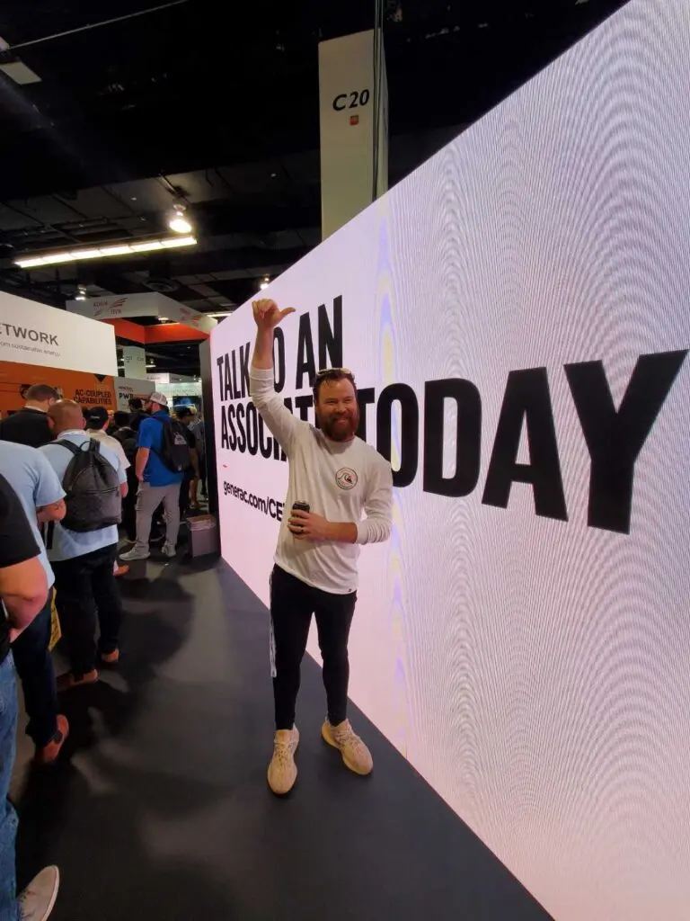 Man holding a drink and giving a thumbs-up while standing against a massive LED video wall that reads 'TALK TO AN ASSOCIATE TODAY' with a trade-show crowd to the left.