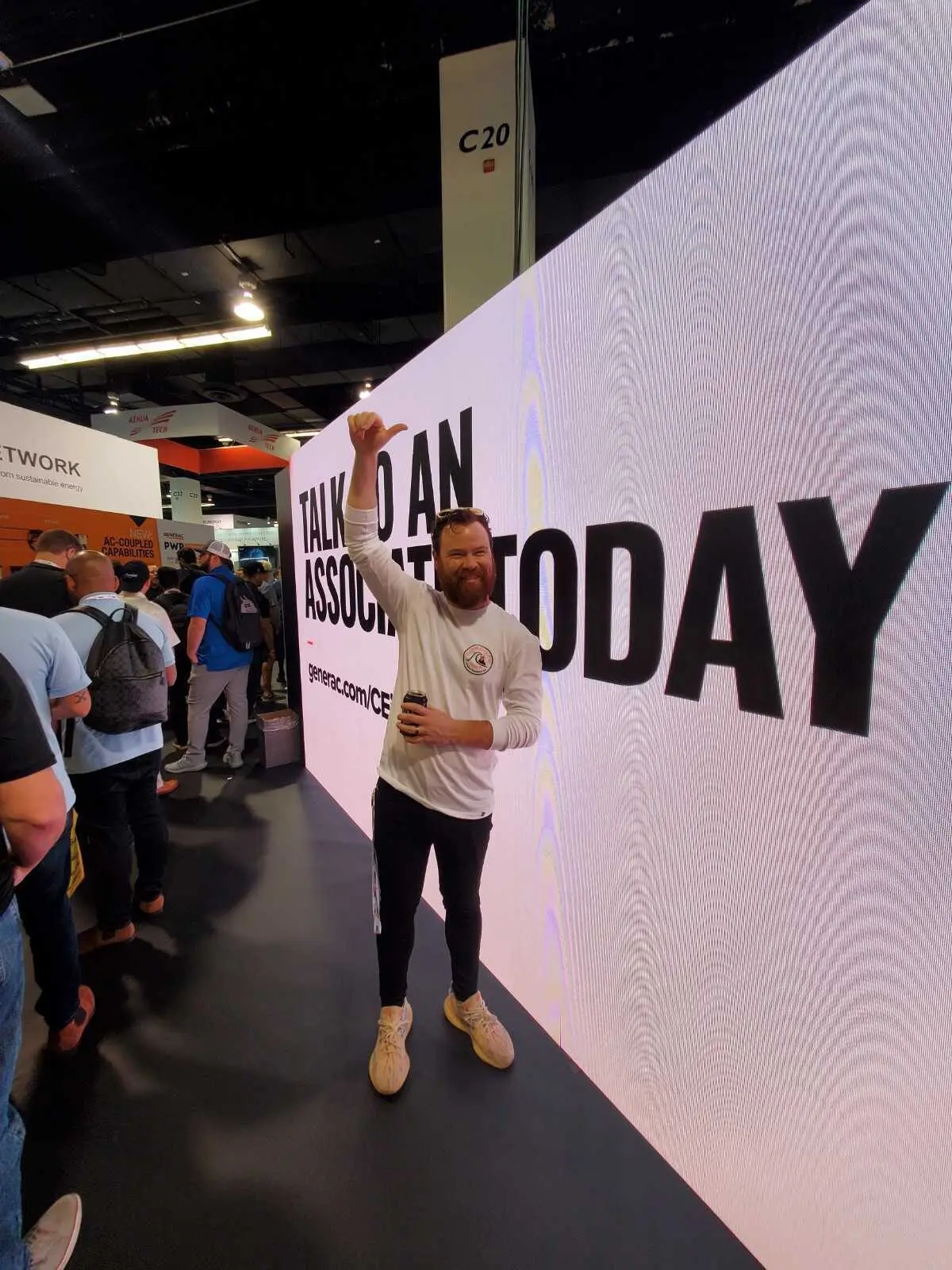 Man holding a drink and giving a thumbs-up while standing against a massive LED video wall that reads 'TALK TO AN ASSOCIATE TODAY' with a trade-show crowd to the left.