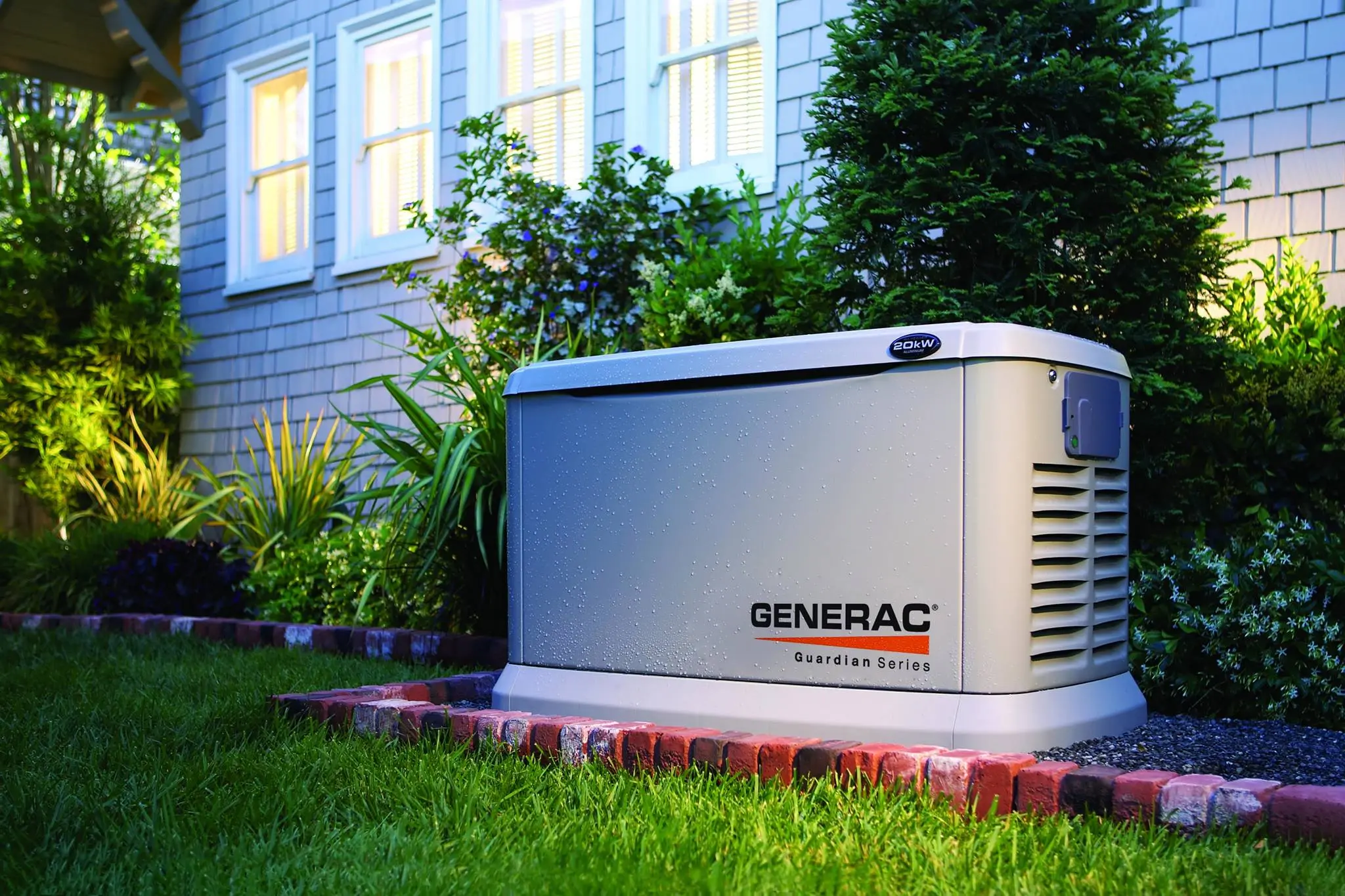 A Generac Guardian Series home standby generator sits on a low brick pad beside a shingled house, backlit by warm interior windows and surrounded by trimmed lawn and shrubs.