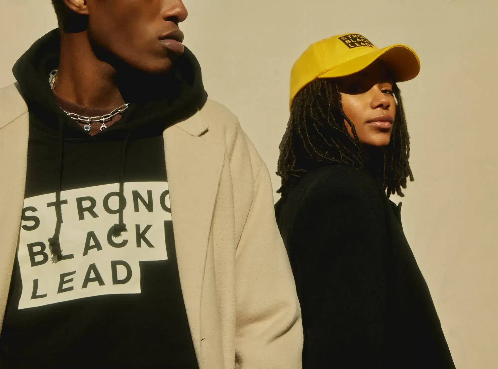 Two people model Strong Black Lead apparel, including a black hoodie with the stepped logo and a yellow cap with a small branded patch.